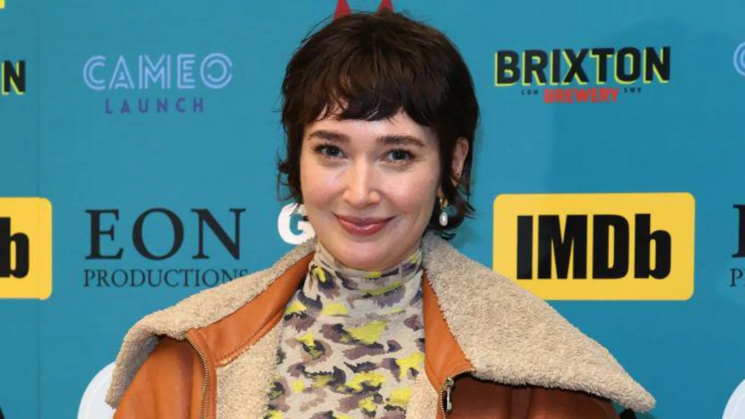  A woman looking at the camera and smiling during a press event. She has short, dark hair with a fringe. She is wearing a tan-coloured leather jacket with a large cream fur collar over a beige animal print turtleneck top. She is standing in front of a blue board which promotes several sponsors' logos.