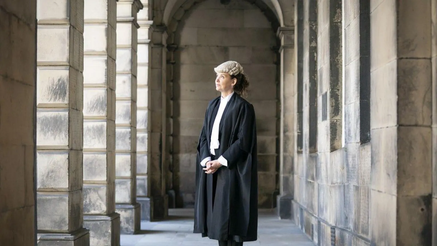  Lord Advocate Dorothy Bain - a woman with dark curly hair tied back behind her head - pictured in the middle distance, standing with her hands clasped, between the stone pillars of Edinburgh's Court of Session, dressed in the traditional outfit of an advocate - long black robes over a white shirt, with a lawyer's wig