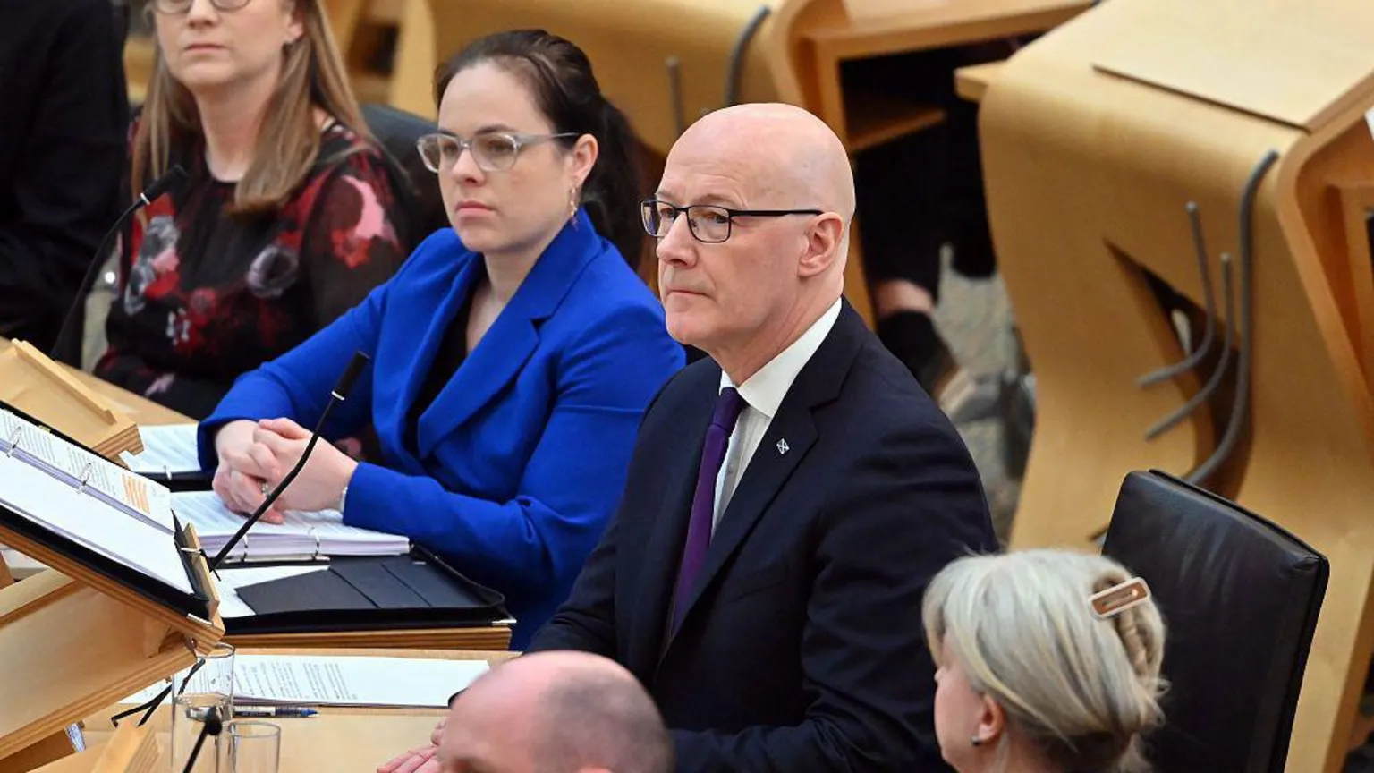  The government front bench at Holyrood - with first minister John Swinney in the centre, a bald man with glasses in a dark suit and purple tie. To his left is his deputy, Kate Forbes, a woman with dark hair tied back behind her head, glasses, and a royal blue suit.