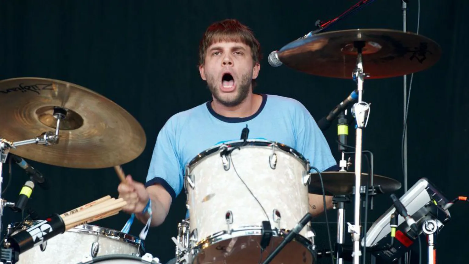  A man in a blue T-shirt with a beard and brown hair plays a white drum with cymbals beside it