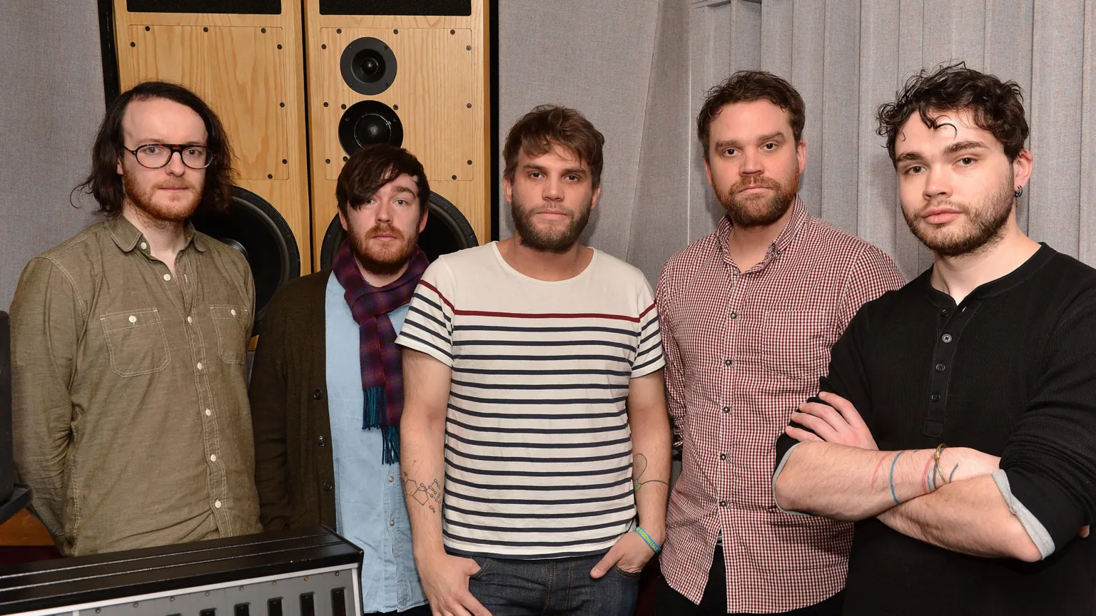 5 men face the camera in a line. closest on the rght has his arms folded and has wavy hair. far left has glasses, thin hair to his shoulders and a larger forehead. to his left the man has a scarf and baby shirt on, his hair flops to one side and is darker.