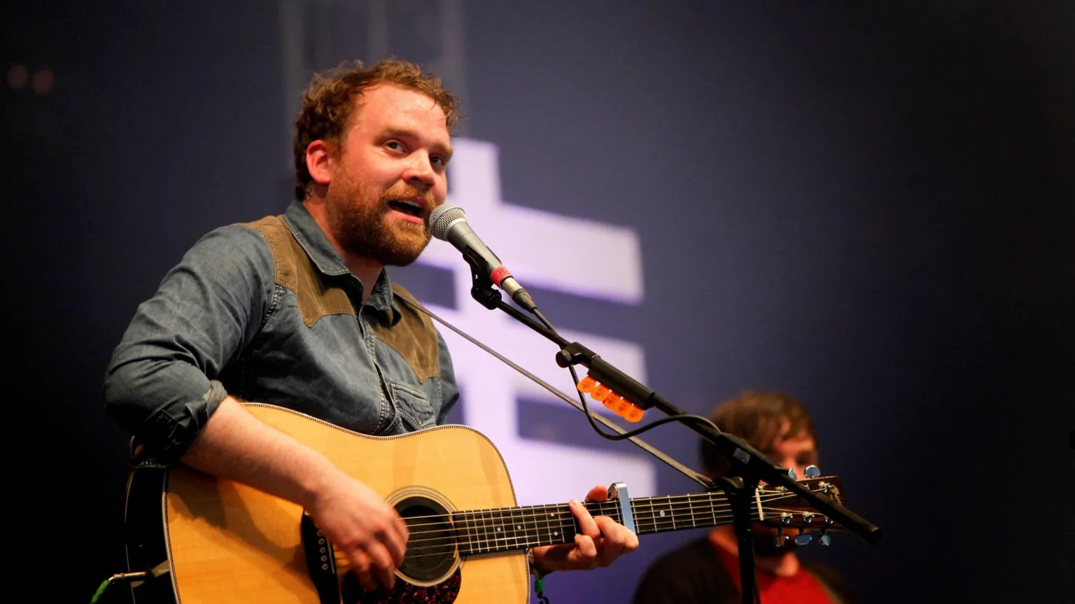A man in a denim shirt is on stage and singing into a microphone on a stand. he is also playing an acoustic guitar.