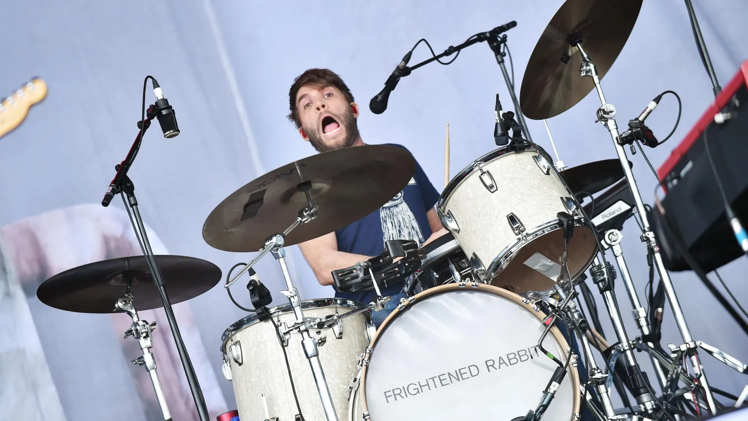 A man sits behind a drum set on a stage. He had red ear plugs in his ears. shaggy beard and hair. he also has his mouth open as if he was singing along with a rock song.