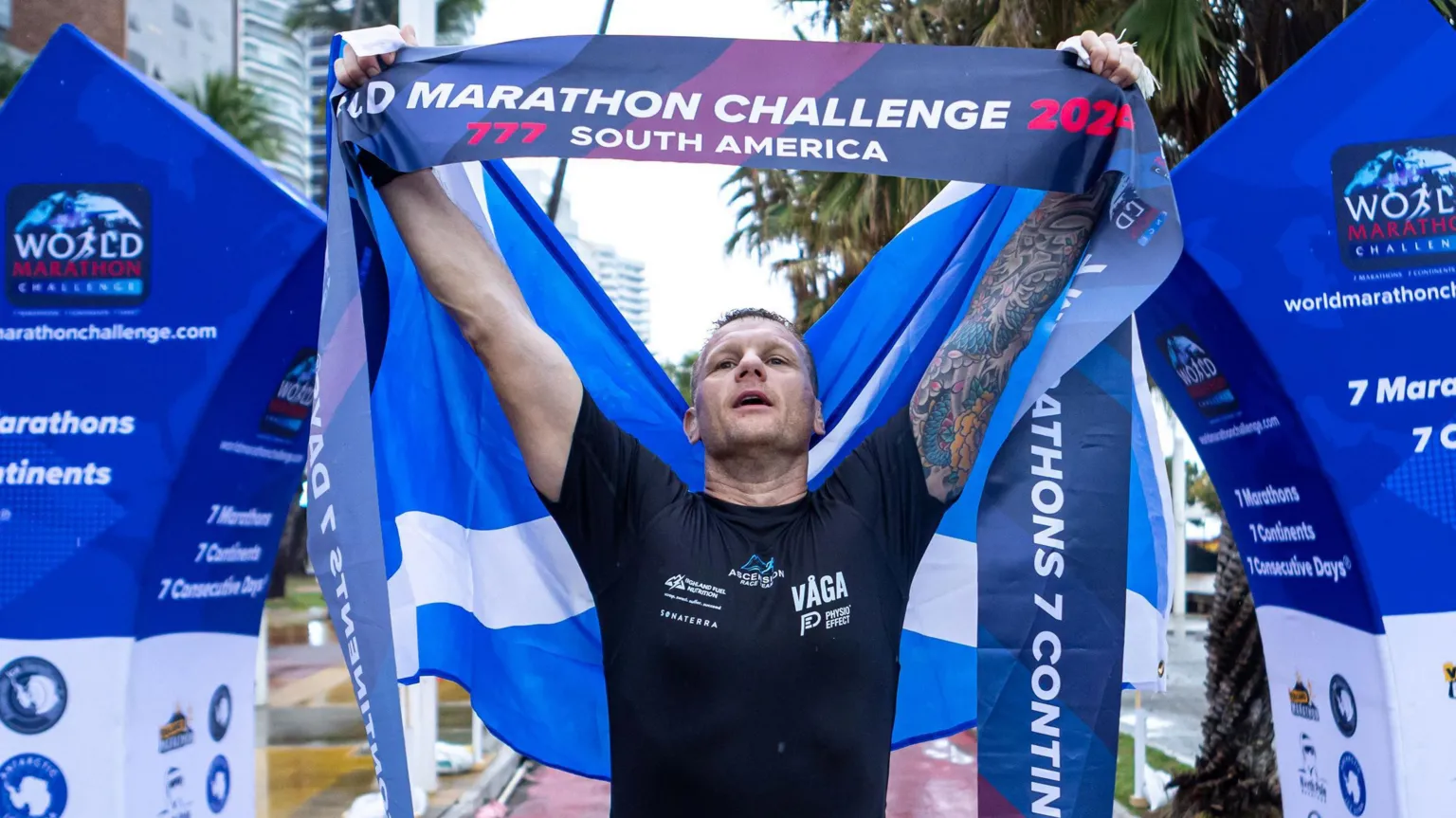 World Marathon Challenge Bobby after crossing the finish line after his penultimate marathon in Brazil. His arms are raised in triumph and he is holding the finishing line tape which reads: 