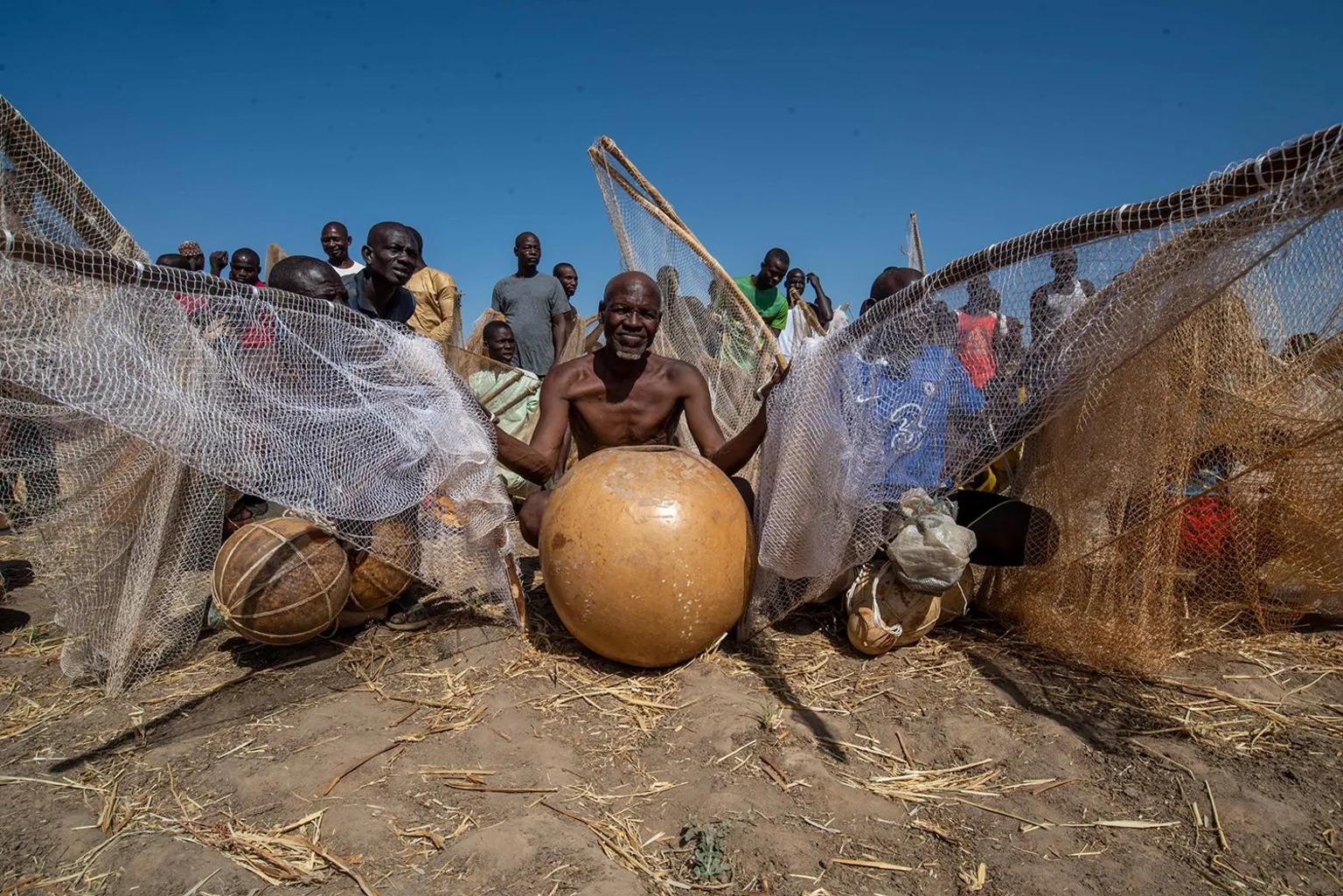 Sani Maikatanga A bare-chested fisherman sits with his gourd and nets on dusty ground. 