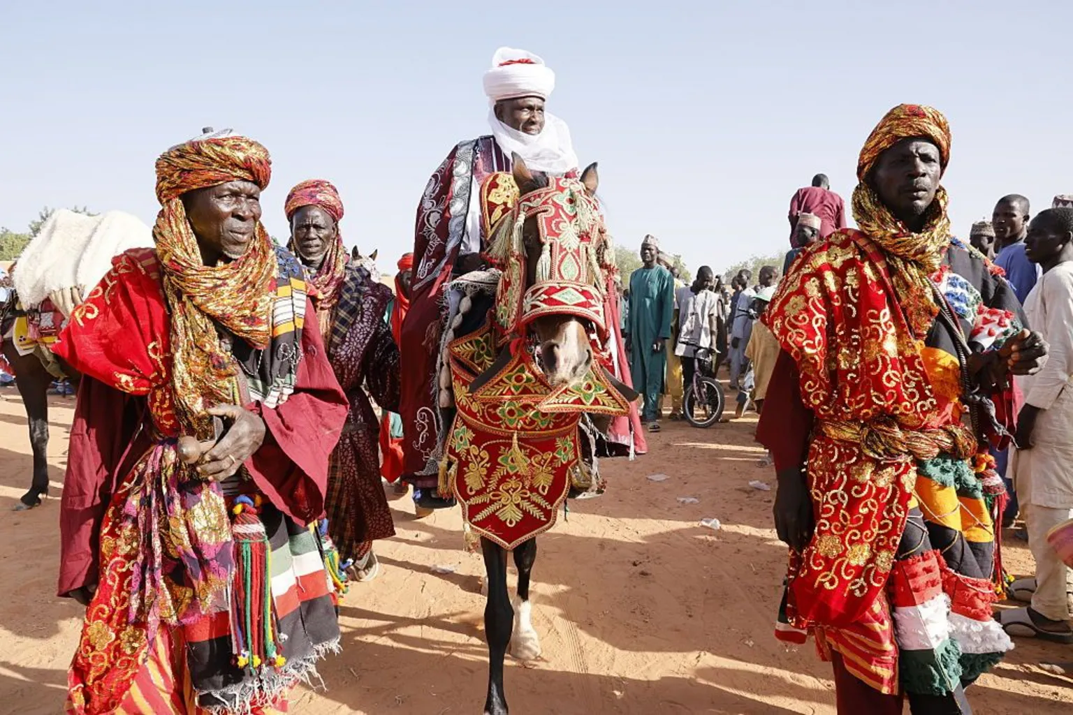 Anadolu via A man in traditional attire and turban is on a horse which is dressed in embroidered red cloth. Two men, in matching clothes are in front leading the procession.