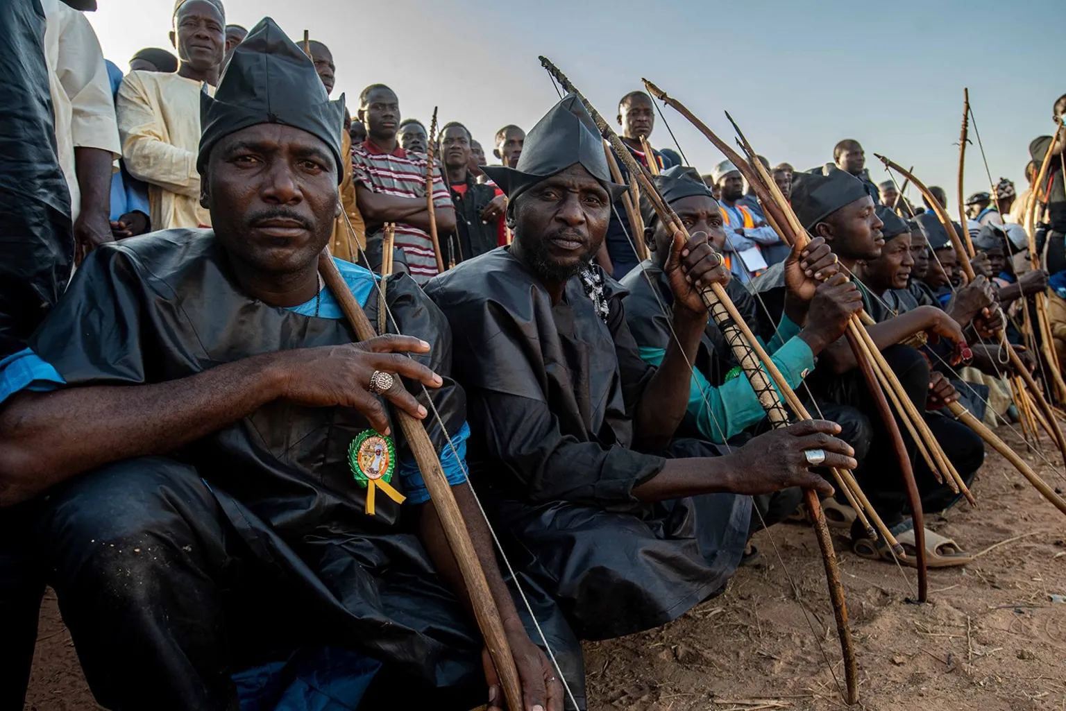 Sani Maikatanga A side view of a row of archers in dark blue attire and holding their bows. They are crouching on dusty ground. 