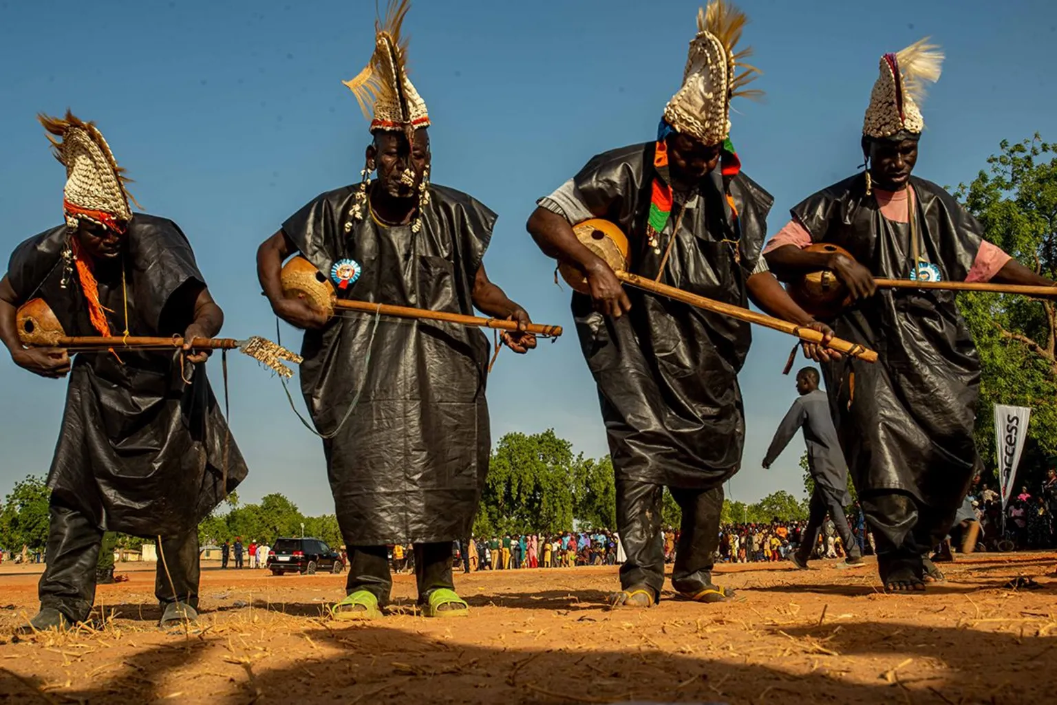 Sani Maikatanga Four musicians in a row in dark attire and a traditional shell covered hat strum guitar like instruments. 