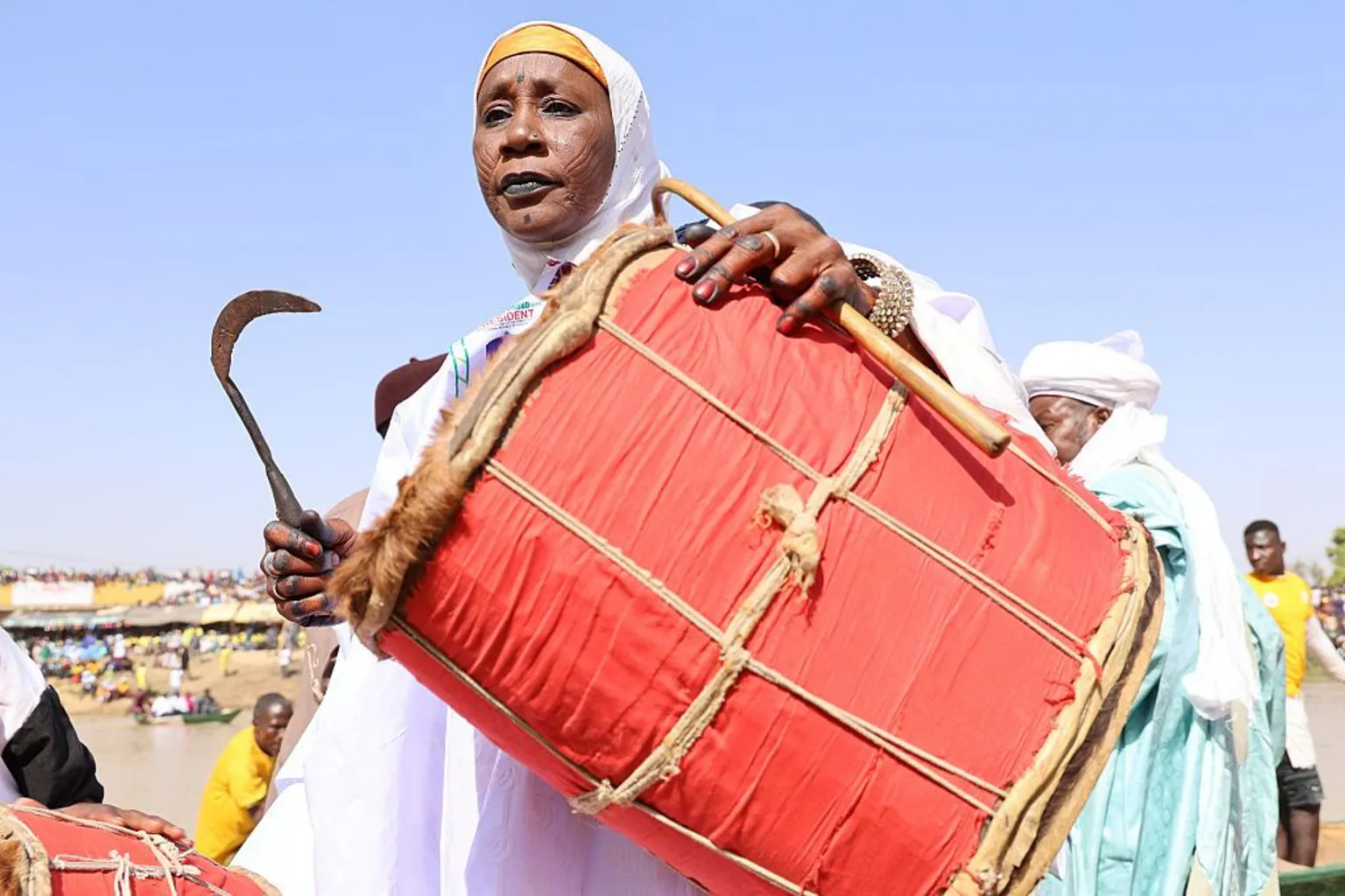 Anadolu via A woman, in a white garment and head covering, cradles a large red drum and holds a sickle-like beater in her right hand.