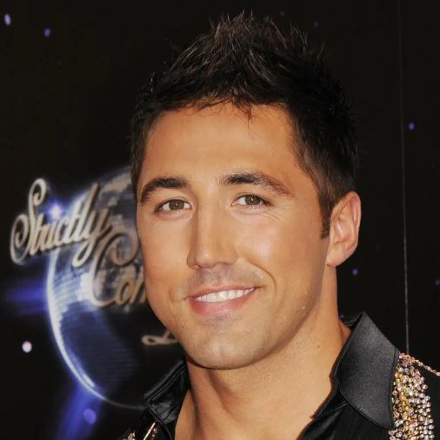  Gavin Henson on Strictly Come Dancing. He looks at the camera and smiles. He has a black shirt on with sequins around the collar and has his gelled hair. Behind him is a Strictly logo.