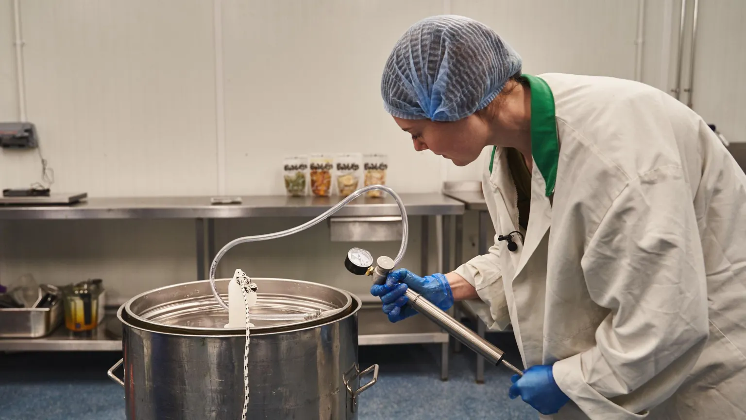 Bwblin A woman in a laboratory white coat checks a pressure gauge attached to a steel vat inside a food making facility. She is wearing a blue hairnet and blue plastic gloves.