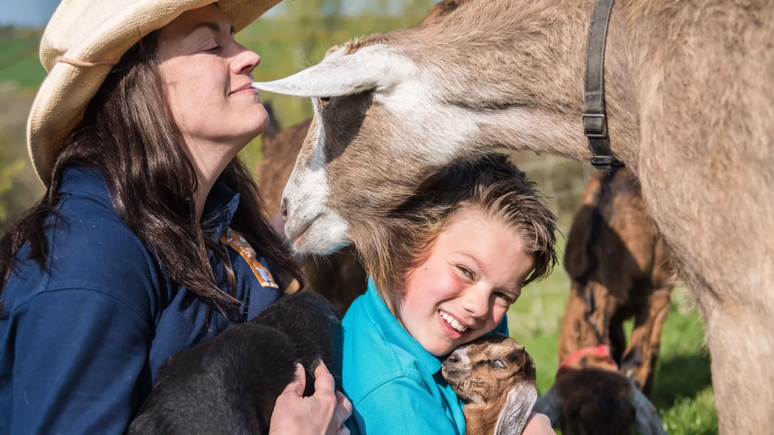Chuckling Goat Shann Jones and son Benji sit on the grass. A large brown goat sniffs near Shann's face while Benji holds a small brown baby goat with long floppy ears. Shann wears a wide brimmed hat, while Benji wears a bright blue fleece.