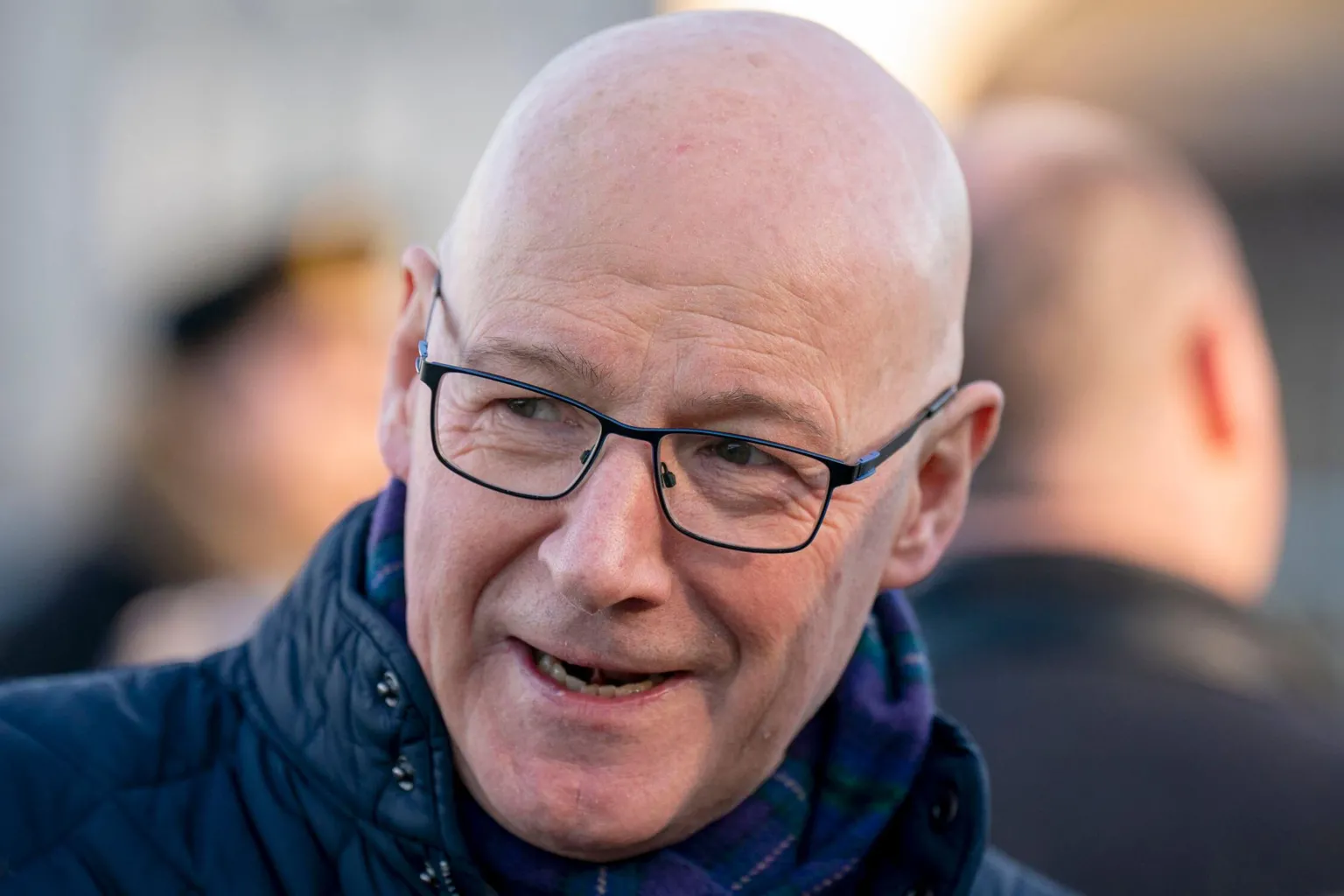  John Swinney is a bald man with dark glasses wearing a padded navy jacket. Two blurred heads are in the background and Swinney smiles mid-convo while looking off to the side 
