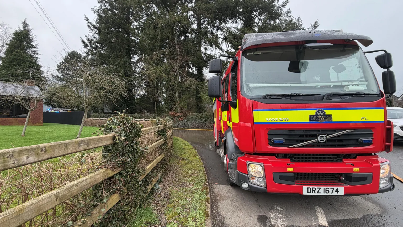 A red, yellow and black fire engine is parked beside a wood fence. Trees are in the background. 