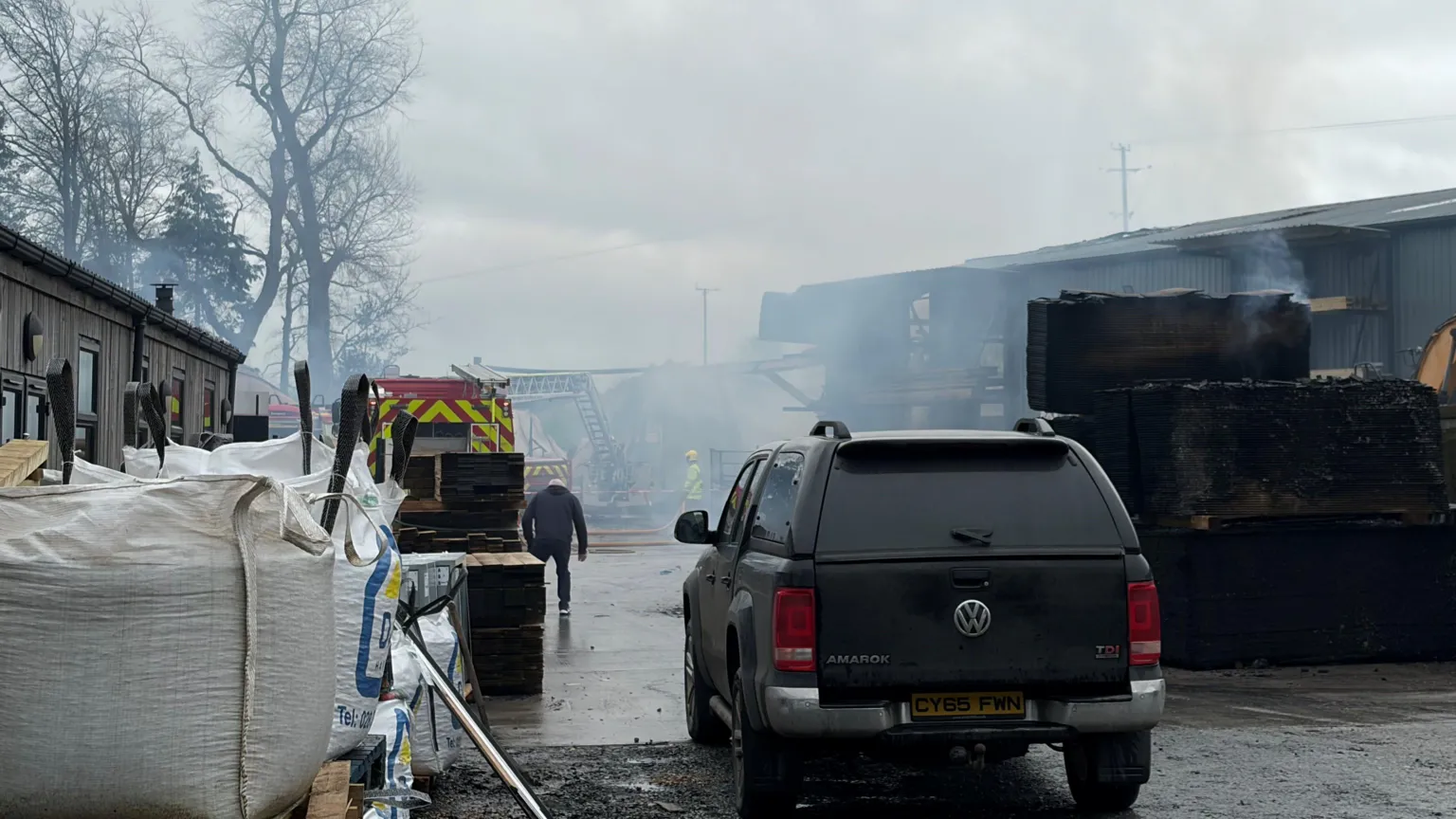 Smoke is in the air beside a building. A fire engine is parked nearby. A car is parked in the foreground. 