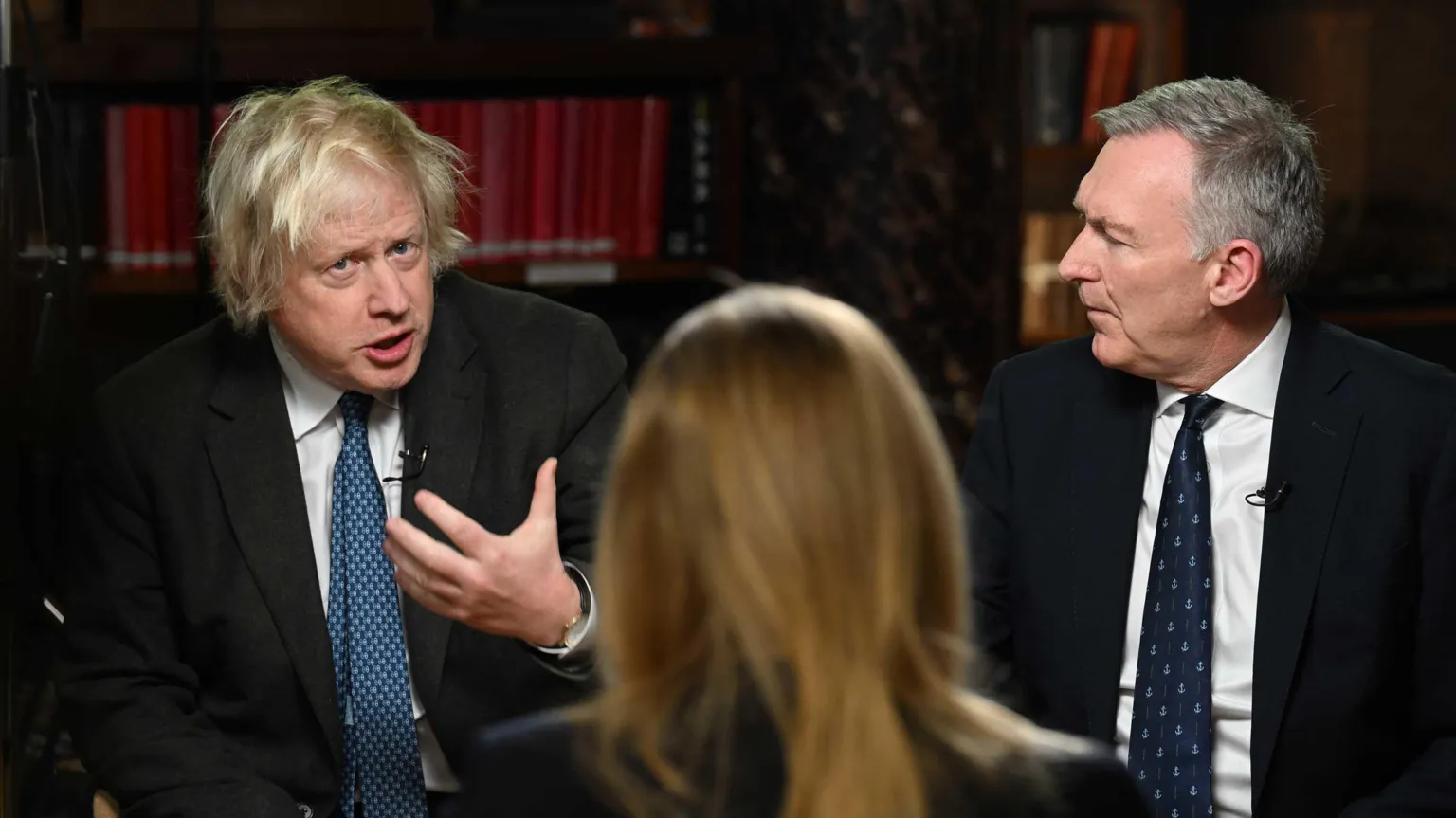 Former prime minister Boris Johnson gesticulates while speaking . A suited man with greying hair looks at him. In the foreground, out of focus, we see the back of a woman with blonde hair.