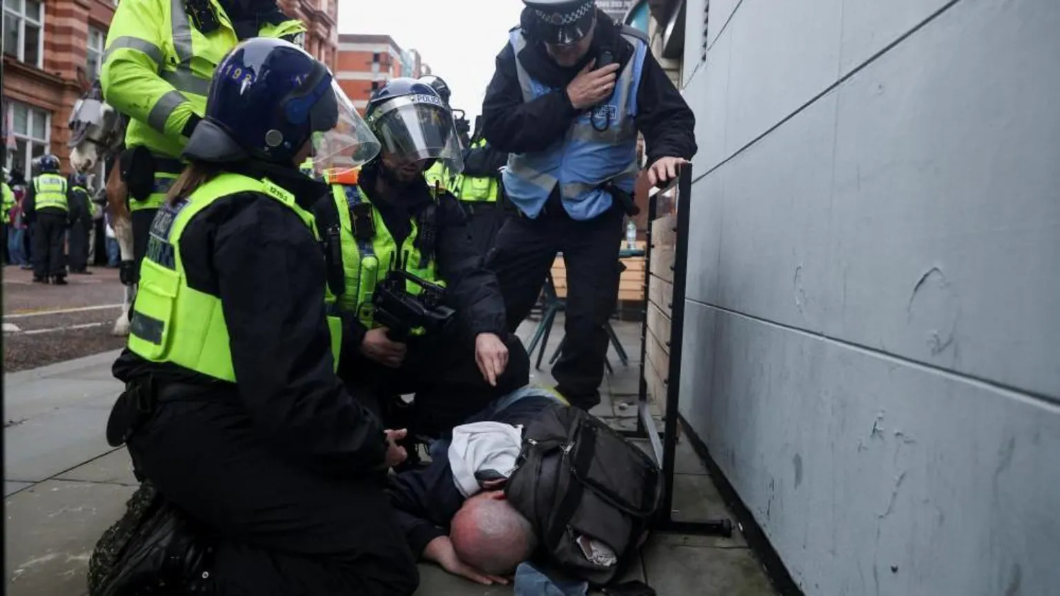  A man is lying down on the pavement with a black rucksack behind him. He is surrounded by police officers wearing helmets