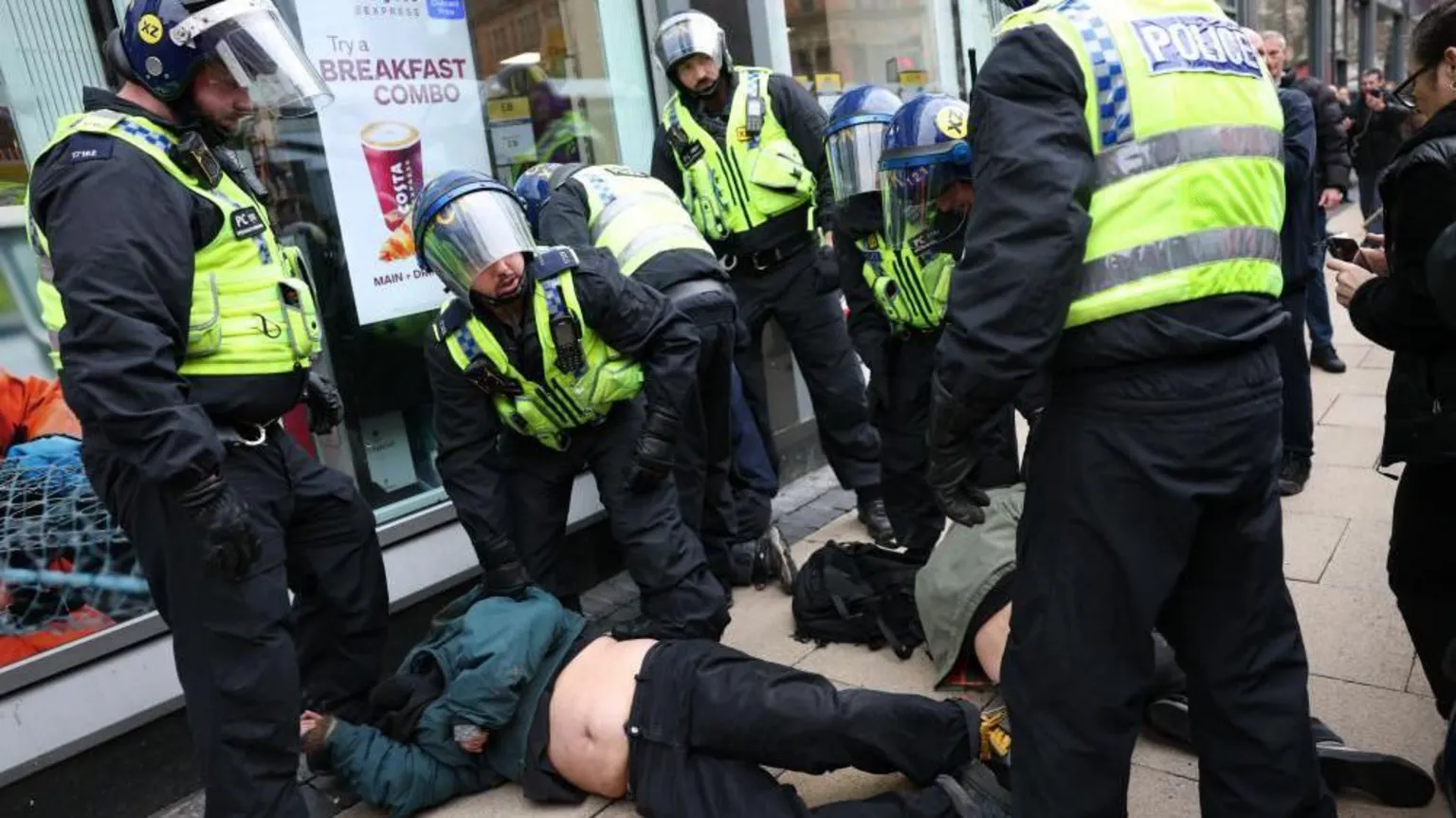 EPA/Shutterstock Two people are lying down on a pavement outside a coffee shop while surrounded by police officers wearing helmets.