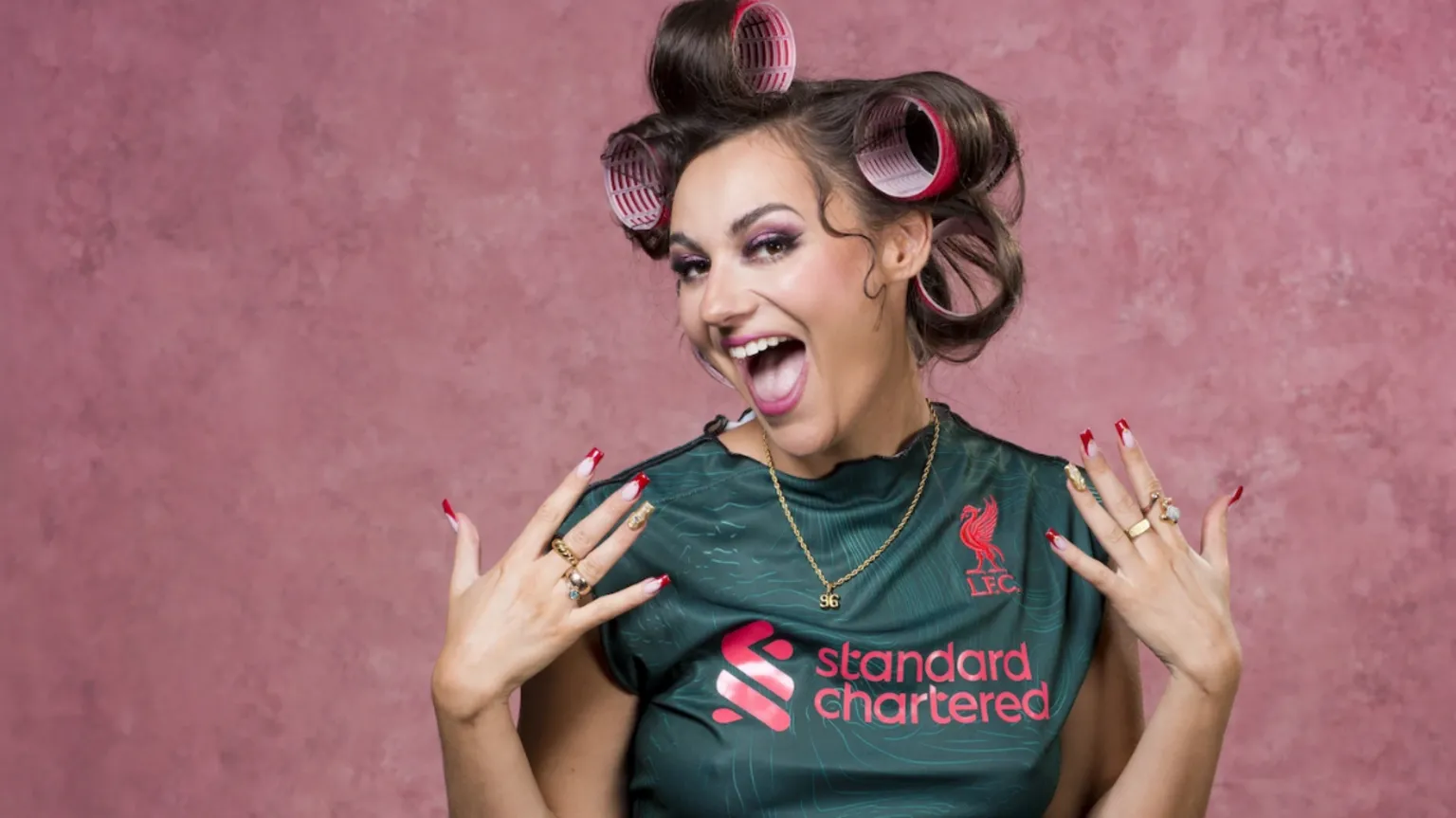 Holly Revell Franks wears rollers in her hair, a Liverpool football kit and has long fake nails. She smiles at the camera with her hands held up. 