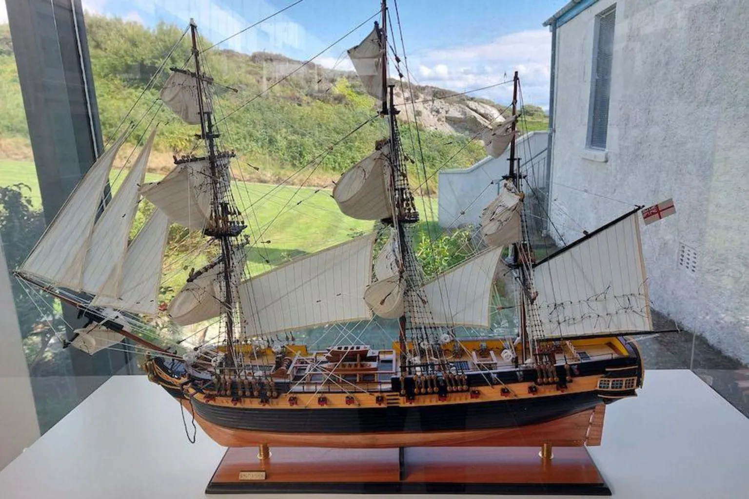 Fort Dunree A side on view of the model replica of the HMS Saldanha on a plinth in a glass box. It is a black and gold wooden sailing ship with numerous sails and lots of rigging. 
