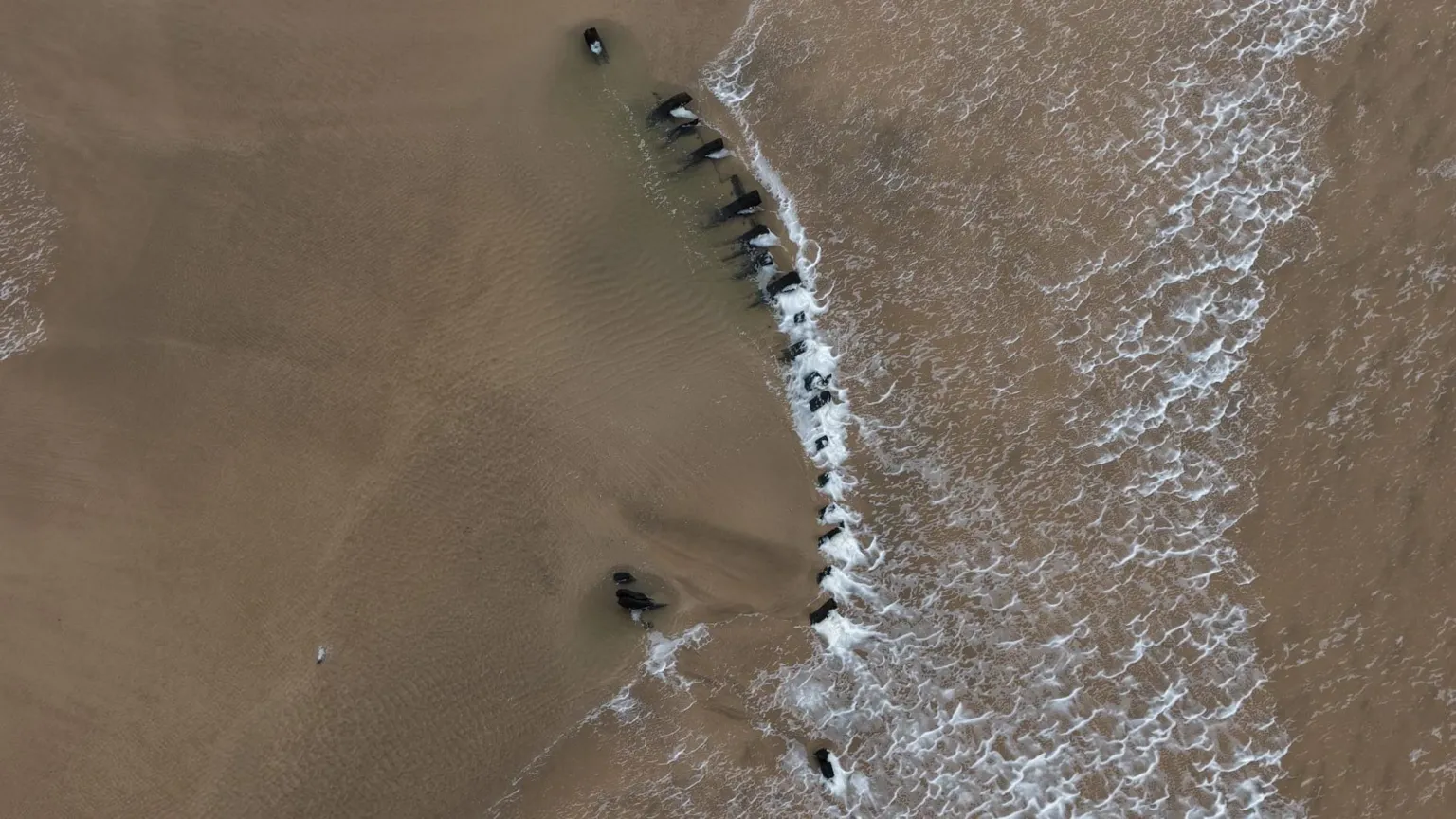 David McGloin Aerial shot of the wooden structure on the beach. A wave passes over a number of wooden pieces that look to be in the shape of a section of a boat.