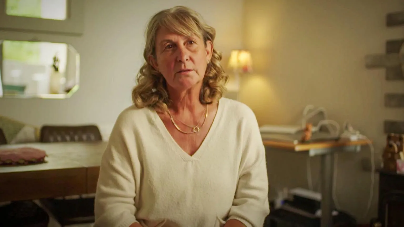 BBC One Wales Photograph of Judith's neighbour, Michelle. She wears a white v neck jumper and has blonde shoulder length short hair. She wears two gold necklaces and looks sadly into the camera. A lamp can be seen illuminating the walls behind her. A dining table is behind her, as well as a desk behind her on the left. 