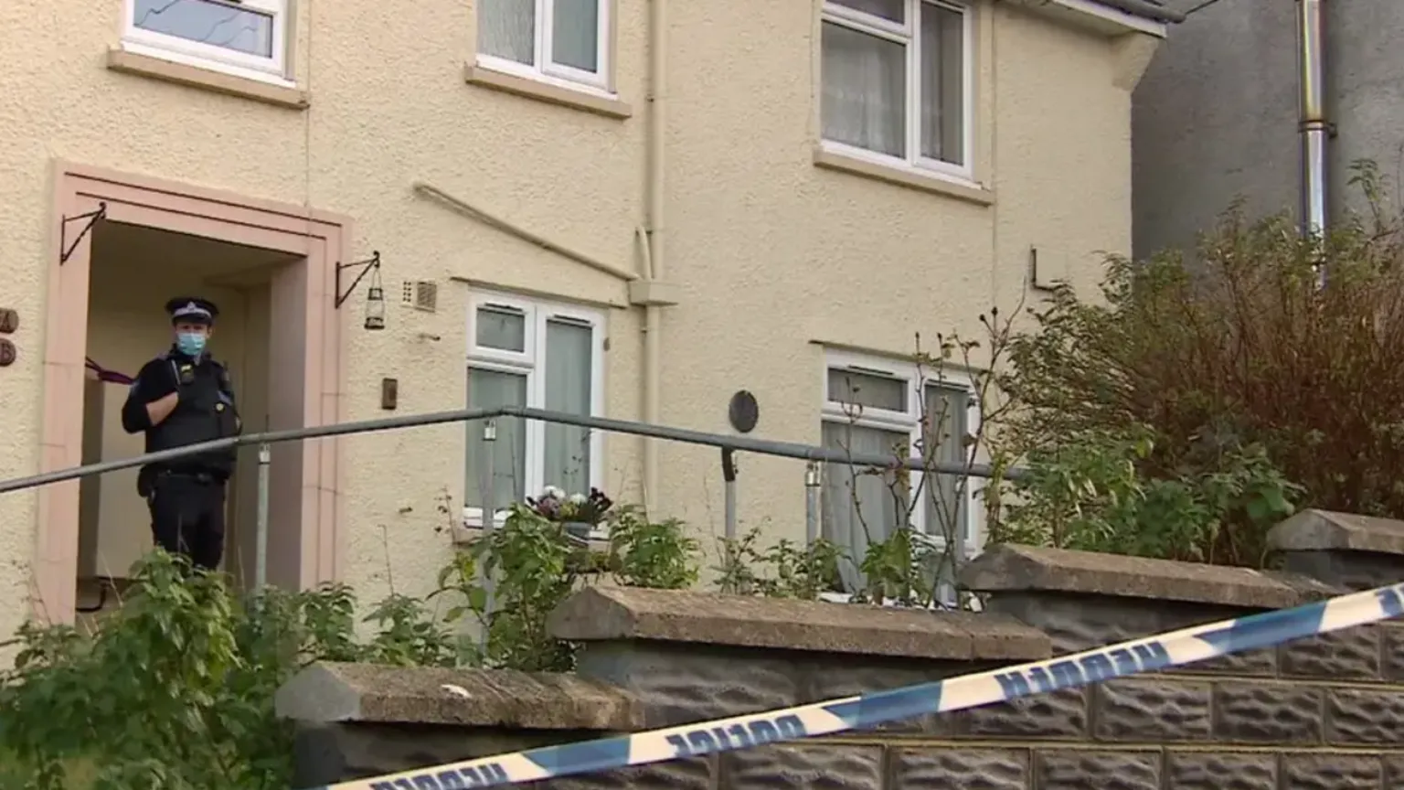 Street photograph of Judith Rhead's home on Market Street, Pembroke Dock. A police officer stands in the door way, wearing a face covering. The door frame is a peach colour, and there are five white windows on the yellow painted building. A support rail is at the forefront of the photograph, just behind a brick inclined wall. Blue and white police tape is seen across the bottom right hand corner. 