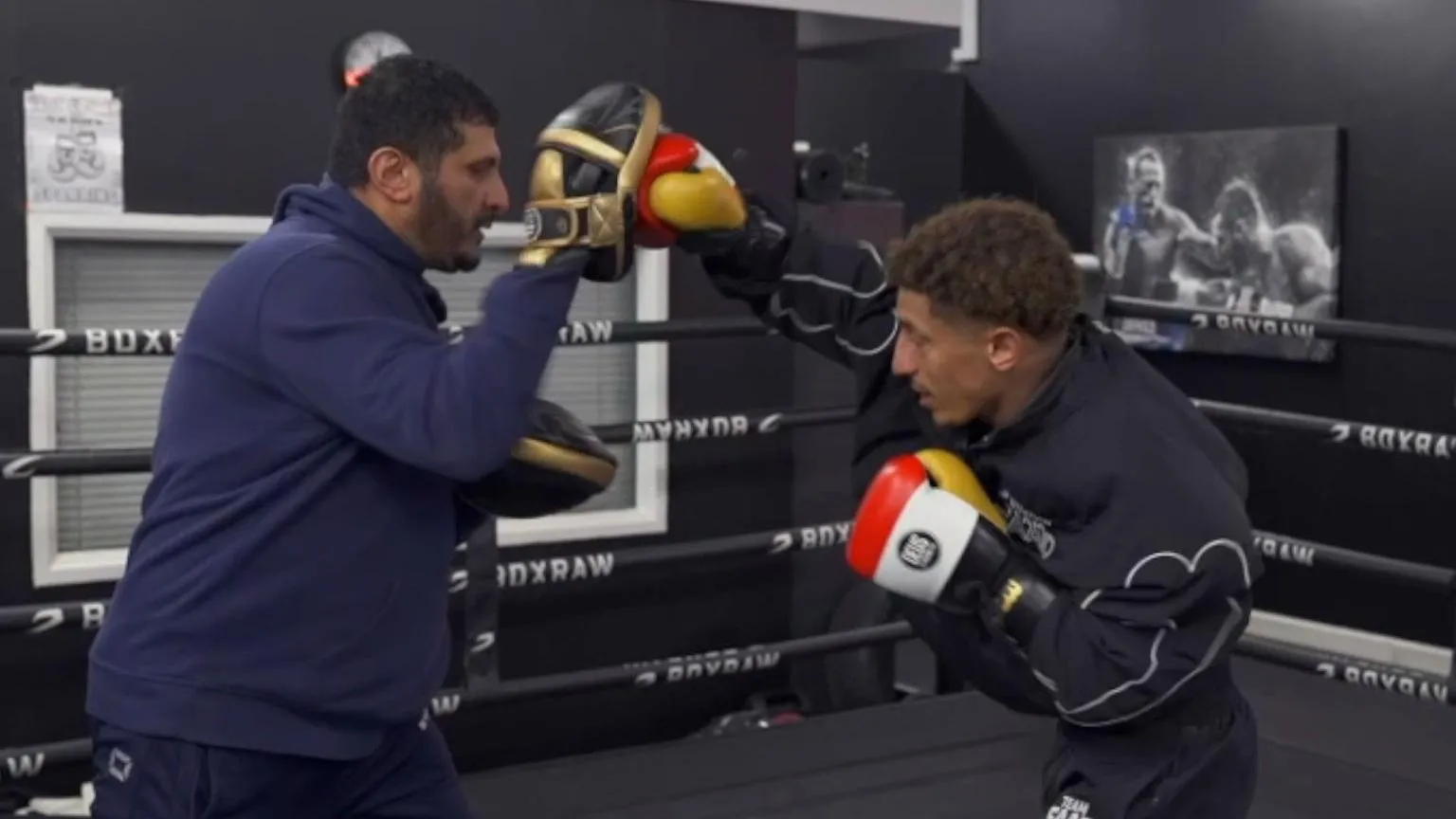 Saad is wearing a black tracksuit and red, white, yellow gloves. He is punching towards gold and black pads that his dad is holding. He is wearing a navy blue tracksuit.