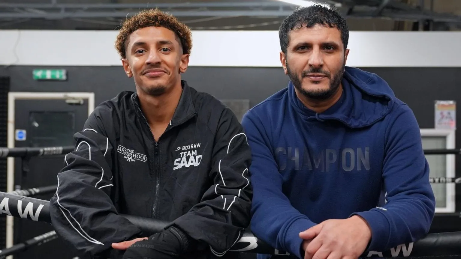 Saad and his dad looking towards the camera and smiling. They both have their hands over a boxing ring bar. His dad is wearing a blue hoodie that says champion on it and Saad is wearing a black jacket.