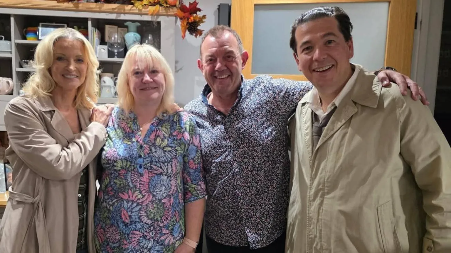 Photograph of Gillian Taylforth (left) Amanda and her husband (centre) and Michael McIntyre (right). Gillian wears a beige trench coat, has blonde short curly hair and holds her one hand on Amanda's shoulder. Amanda has blonde shoulder length hair and wears a blue, pink and green floral shirt. She stands next to her husband who has short brown hair and a navy pattern shirt on. Michael (right) wears a beige coat, white shirt and grey tie. They all smile at the camera. 