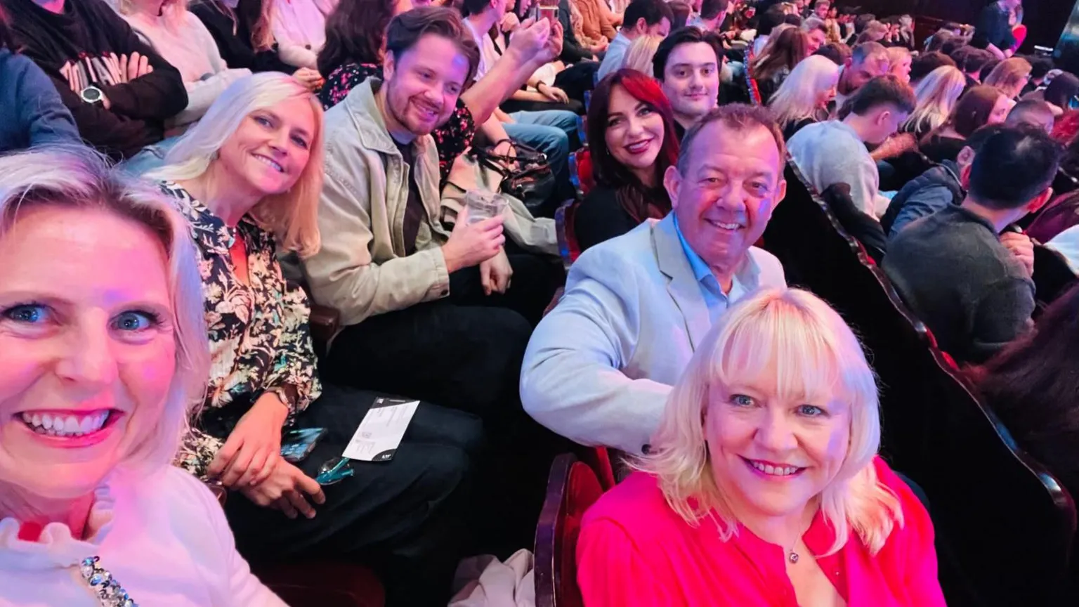 A selfie photograph of Amanda (bottom right) with her husband, son and daughter all along one row in a theatre hall. A lady with blonde hair and blue eyes holds the camera at the forefront, in the bottom left hand corner. In the row behind Amanda is another woman leaning into a young man. They all smile at the camera. 