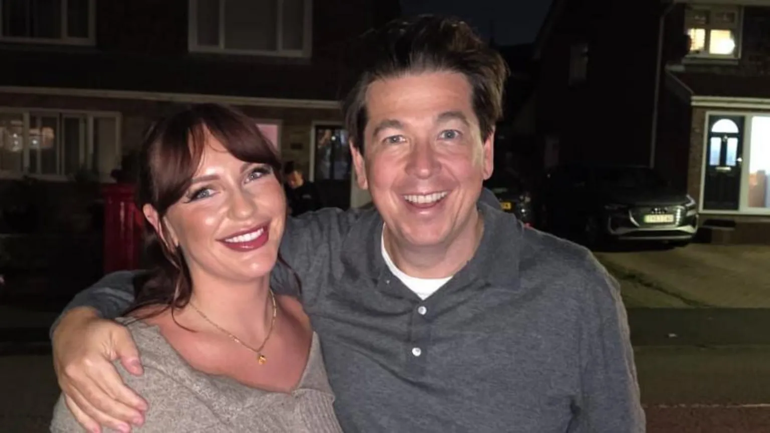 Photograph of Amanda's daughter, Sophie Grey, and Michael McIntyre. Sophie has dark brown hair which is parted into bangs and blue eyes. Michael has blue eyes and brown hair. The pair of them are stood outside at night on a residential road. Michael holds his one arm around her shoulder and they both smile at the camera. 