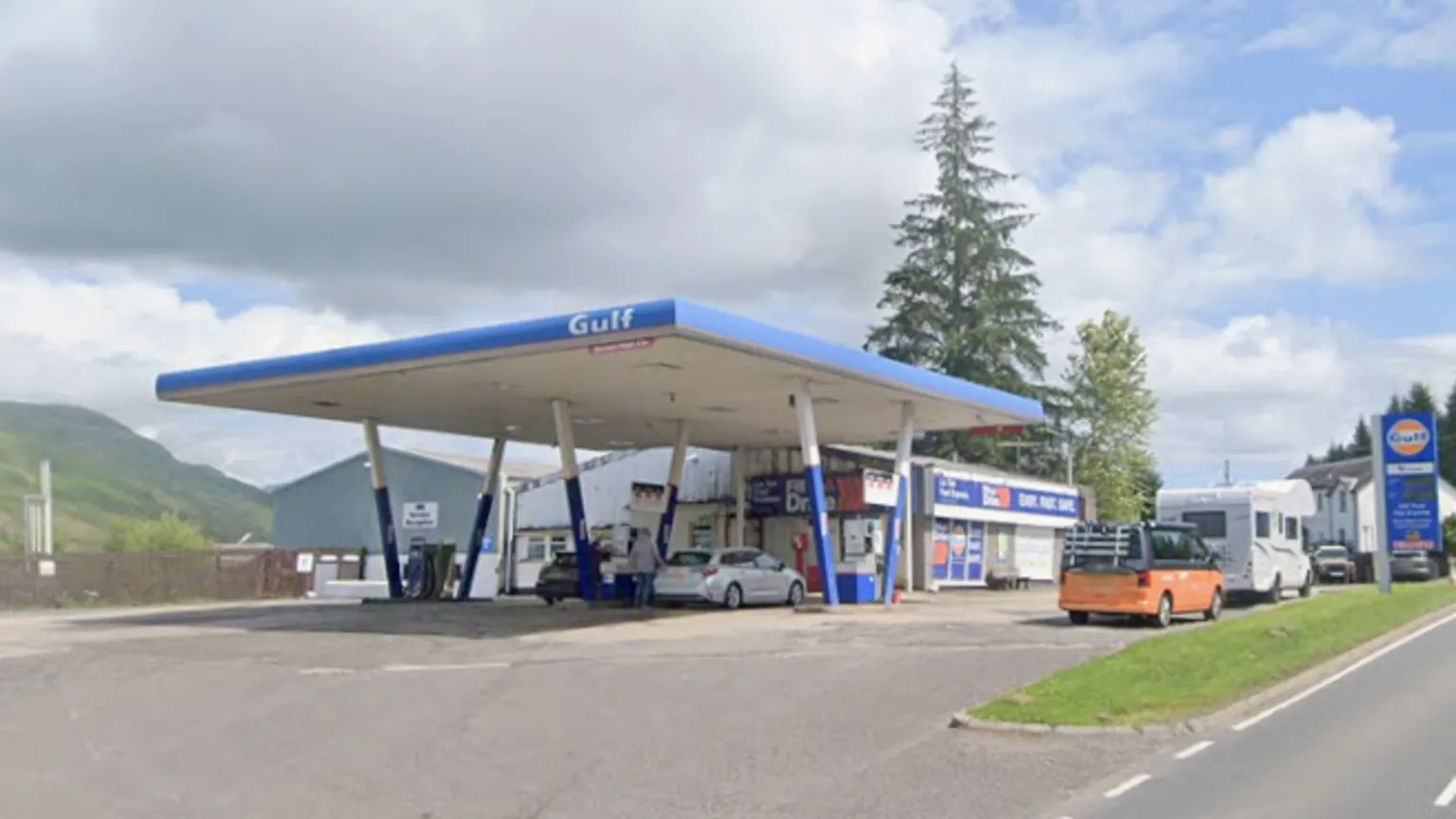 Google Maps Gulf petrol station with a blue canopy over multiple fuel pumps beside a roadside, with cars parked at the forecourt, a shop building behind, green hills in the background, and a partly cloudy sky.