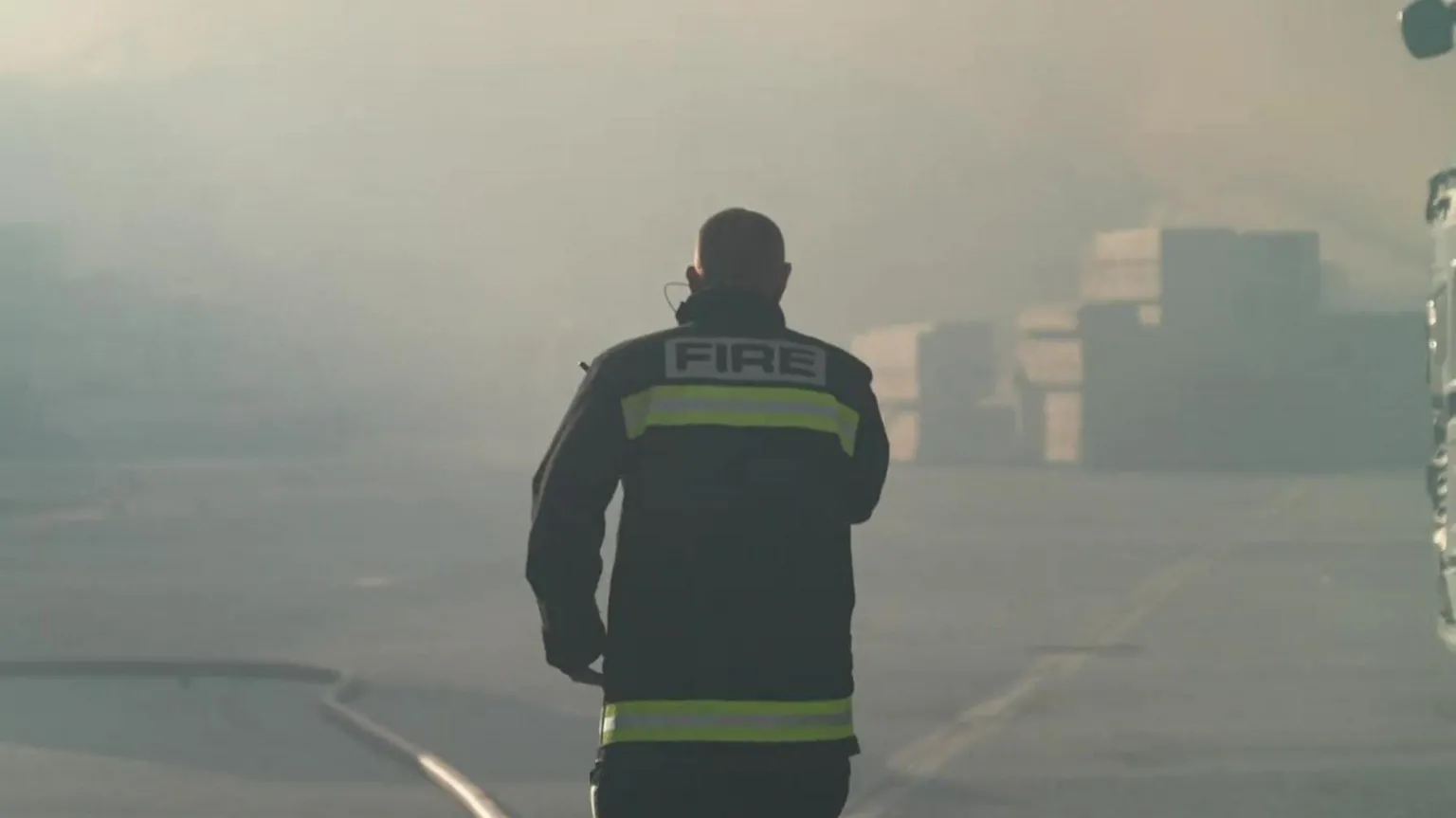 A firefighter walking through thick smoke. He has his back to the camera and is wearing a firefighter's uniform which says 'fire' on the back.