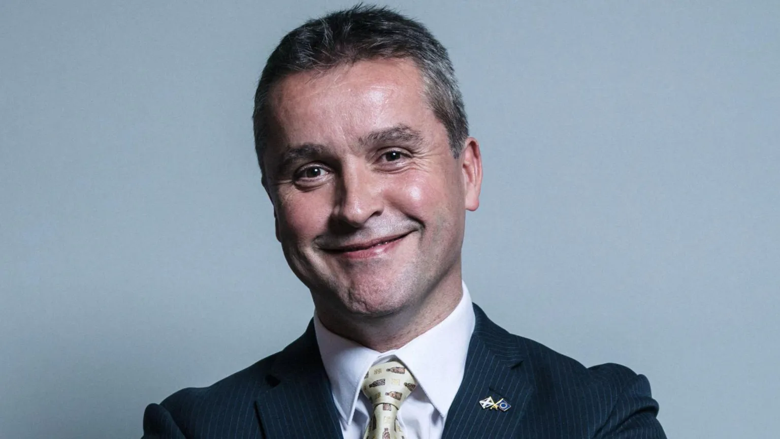  Angus MacNeil has short brown and grey hair and smiles at the camera. He wears a dark pinstriped suit and yellow tie with a white shirt.