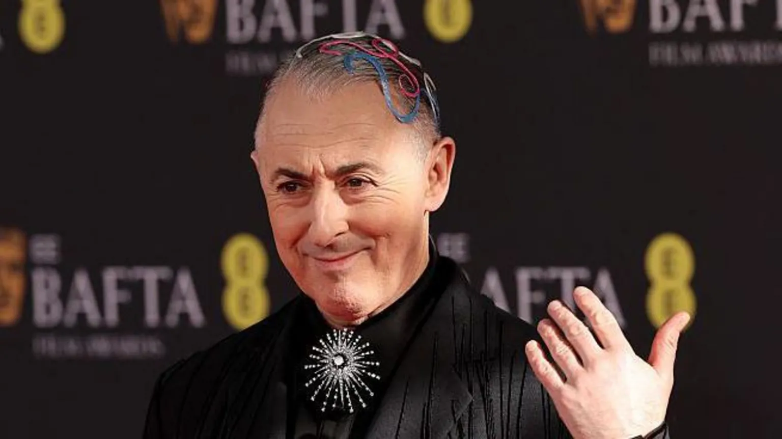  Alan Cumming with colourful coils on his head and a silver, diamond brooch and black jacket and shirt