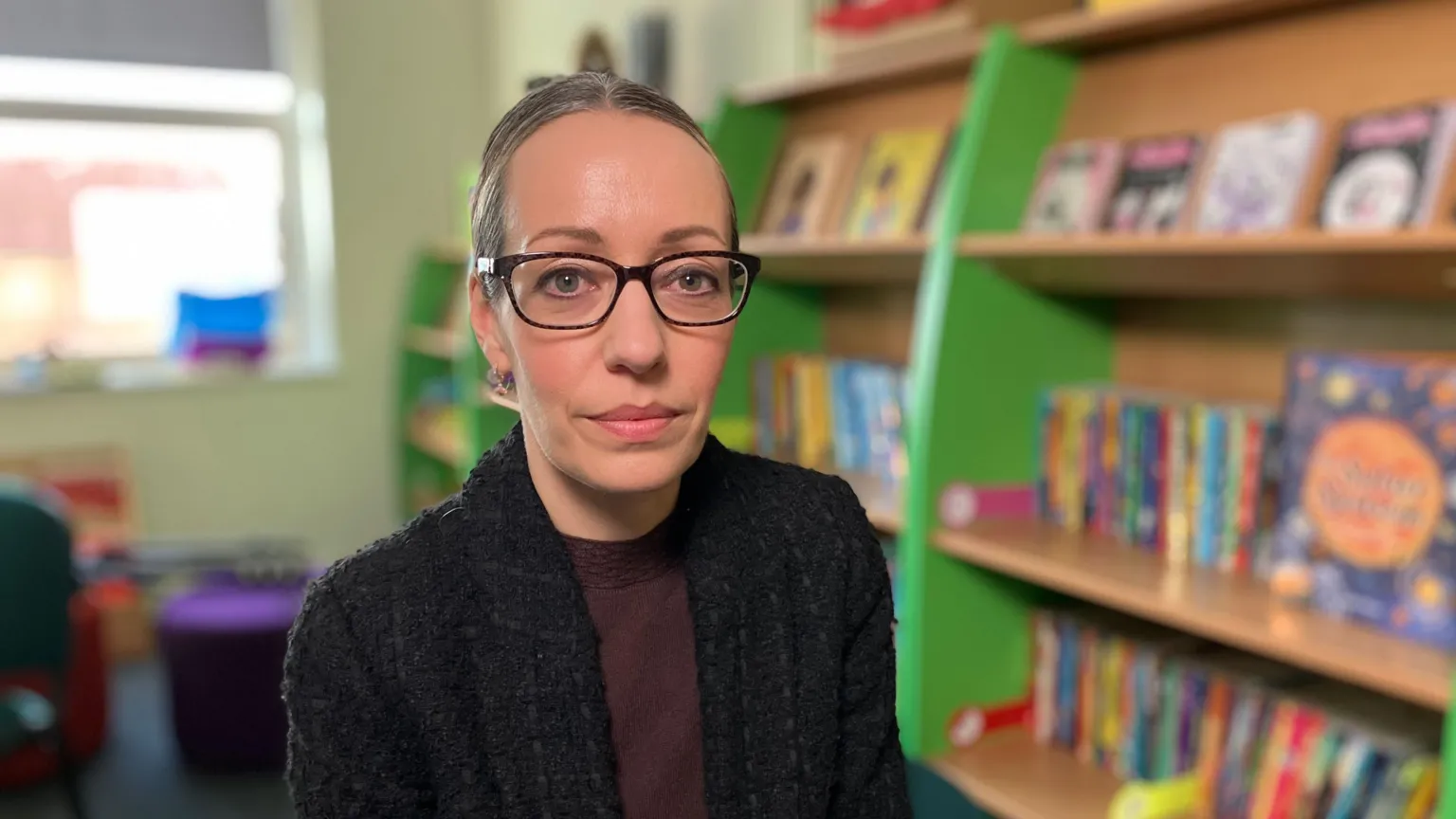 Kate McGough / BBC Claire Robertson is seen wearing a dark jacket in her school's library, surrounded by bookshelves filled with colourful children's books. Stools and cushions are visible in the background.