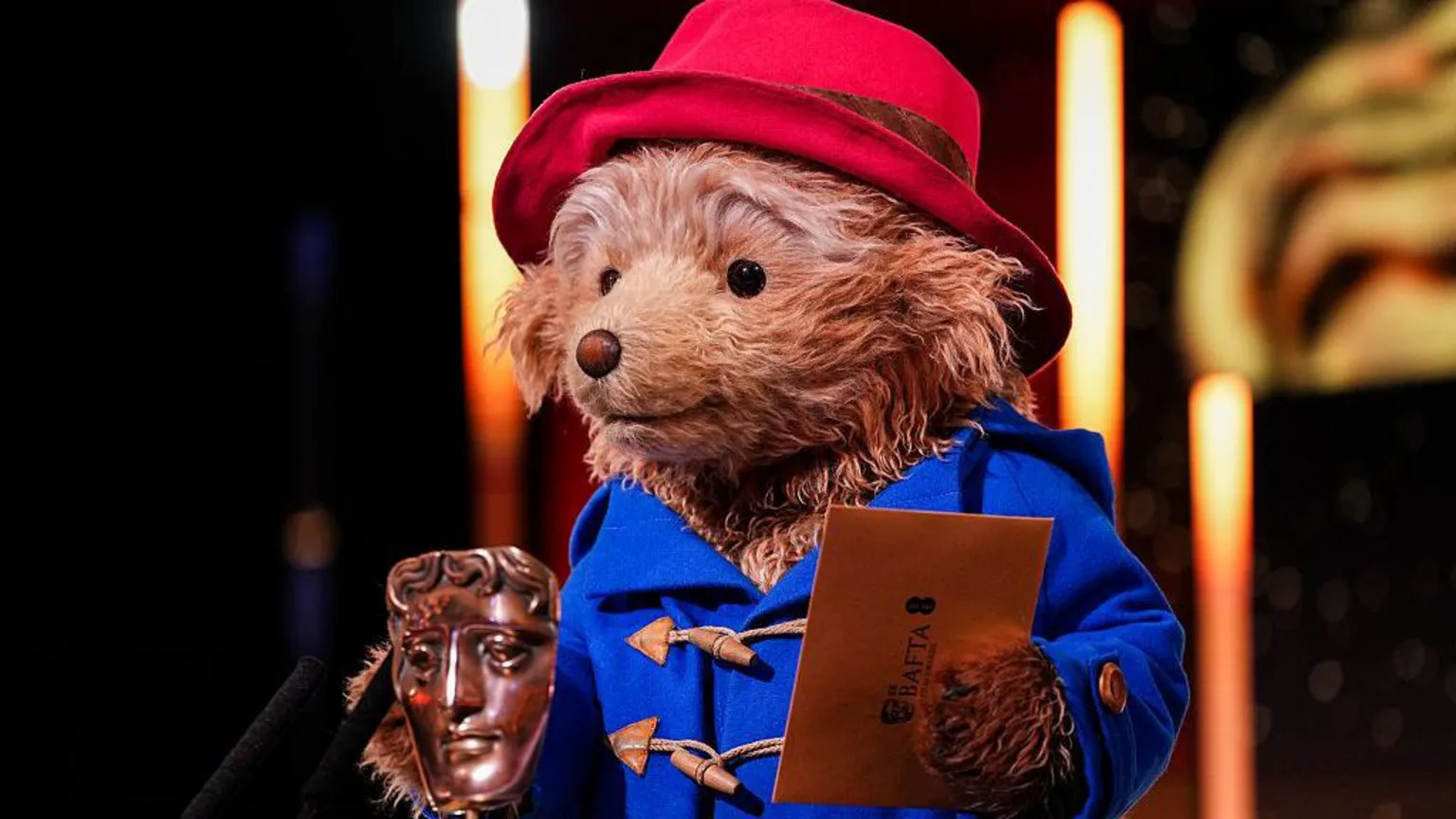 BAFTA via A person in a bear costume holds a Bafta awards envelope in one paw, while there is an award in front of them. They wear a red hat and blue coat 