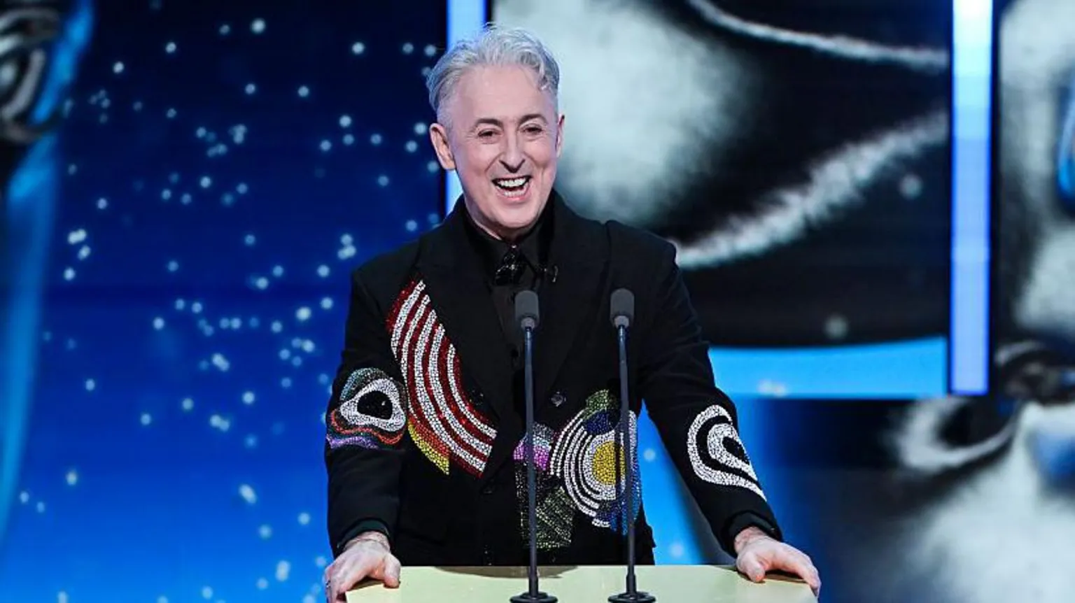  Alan Cumming speaks into two mics that are on a podium which he holds with two hands