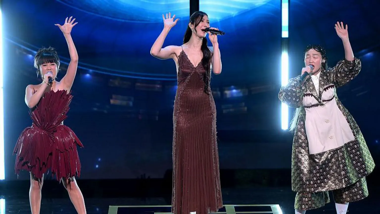  for BAFTA Three women stood slightly apart on a stage in fancy dresses. They each sing into a handheld microphone, while their other hand is raised above their heads