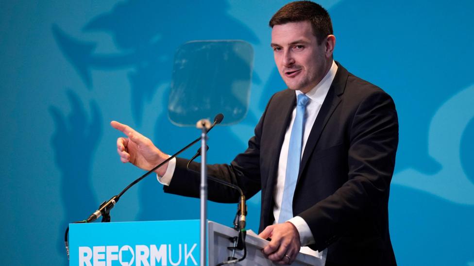 Llun James Evans yn areithio. Mae ganddo wallt brown ac mae'n gwisgo siwt du gyda chrys gwyn a thei glas.