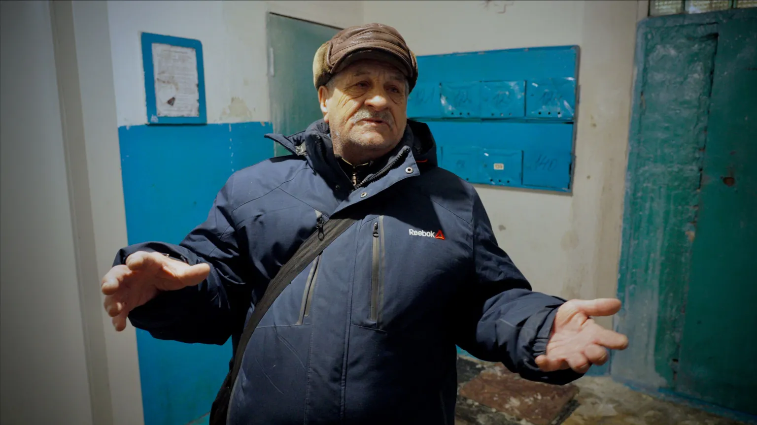 Ivan Pavlovich stands in the hall of an apartment block in Lipetsk. He's wearing a dark blue puffer coat and a brown leather hat.