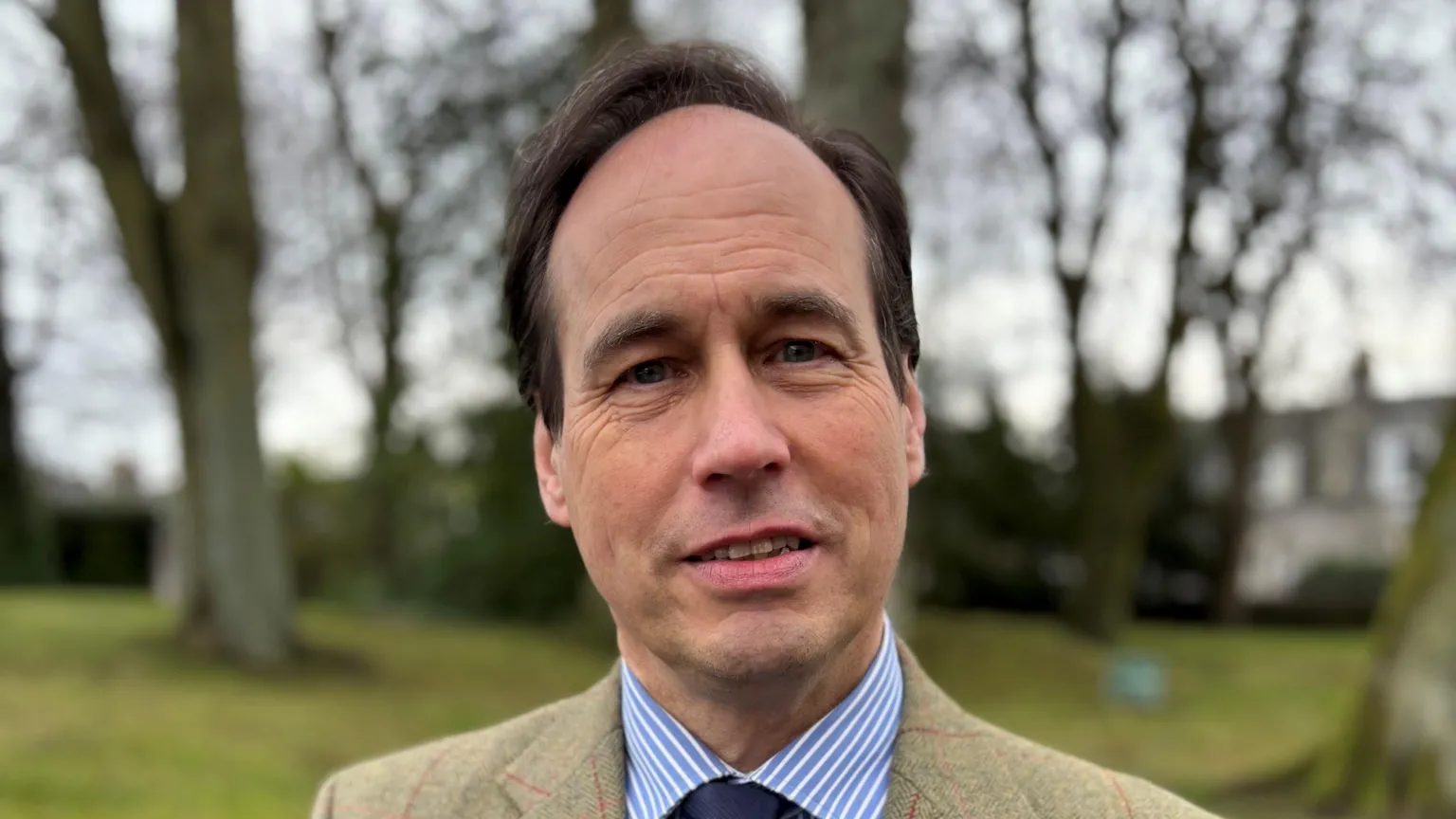 Martin Greig looking straight at the camera. He has dark hair and is wearing a tweed blazer over a blue and white striped shirt and navy blue tie. He is standing in front of a blurred outdoor background with grass and bare trees.