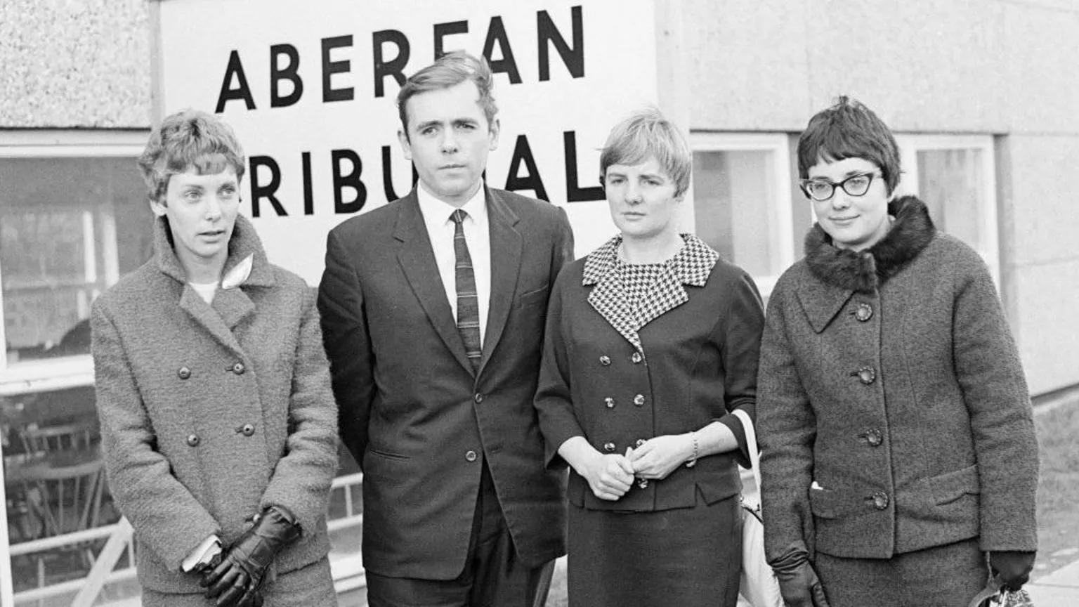  A black and white image from the late 1960s showing four teachers standing in front of a large sign that reads 'Aberfan Tribunal'. The three female and one male teacher are in smart, formal wear. Two of the women have gloves on. Only Mair Morgan, on the right of the image, is wearing glasses. 