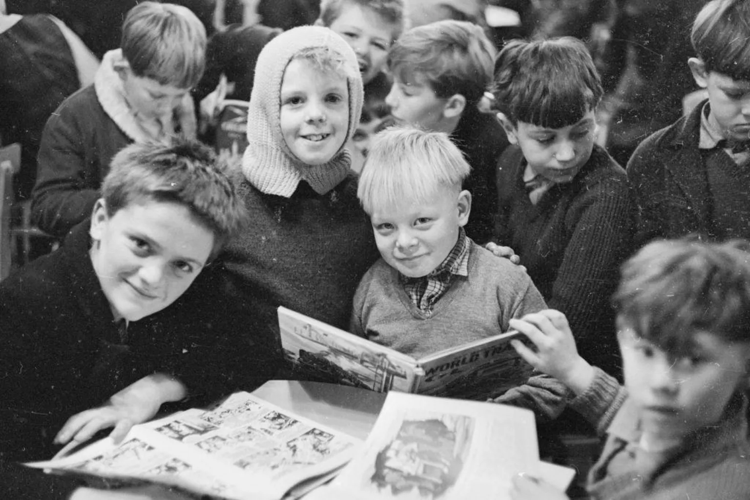  A black-and-white photograph from the late 1960s showing a group of nine boys attending informal school lessons in Aberfan. The boys in the foreground are holding comic books, while one of the group has a light-coloured balaclava on his head. All of the boys are wearing school jumpers, and three of the group are smiling at the camera. 