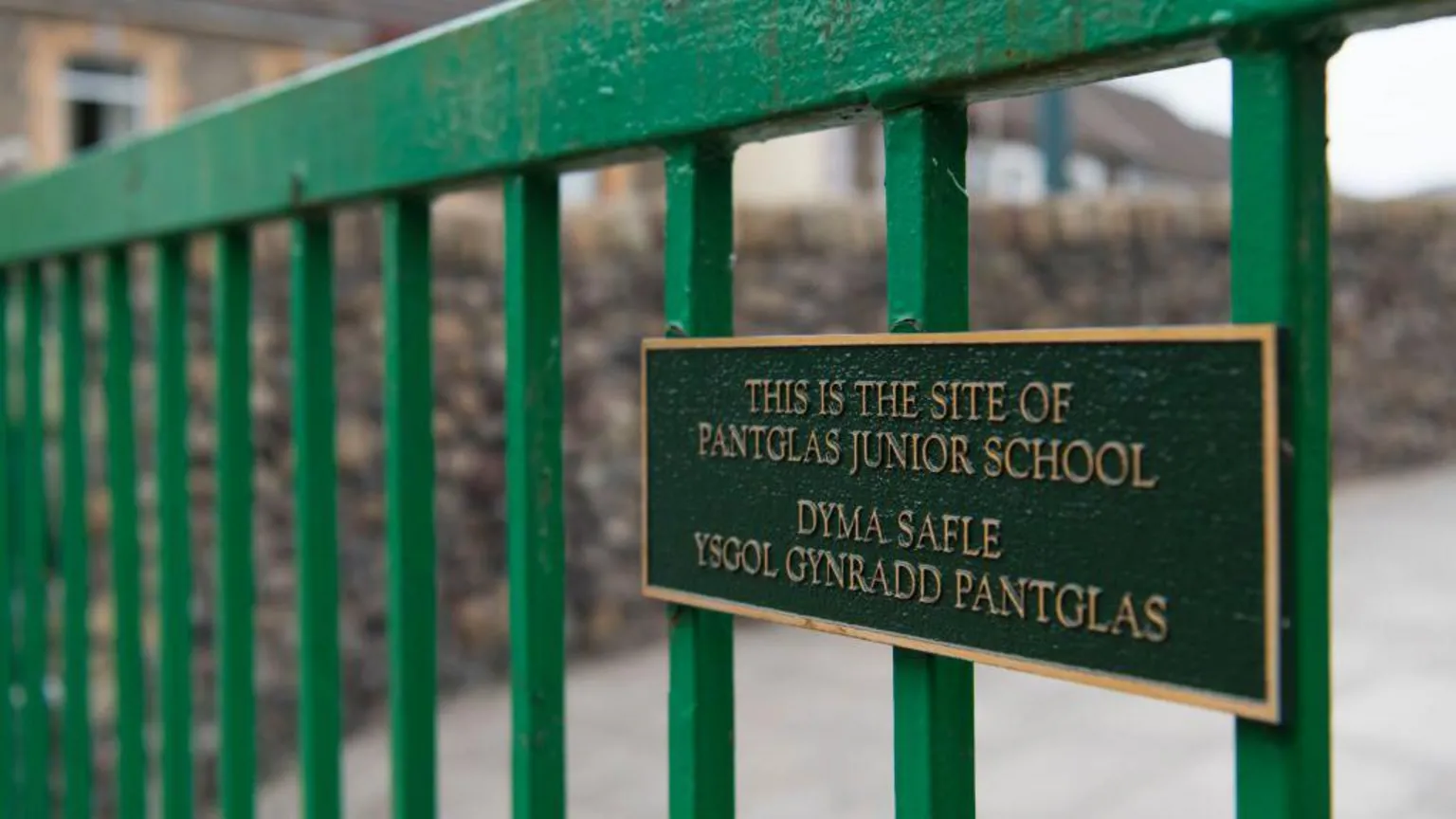  A close-up image of green railings and a plaque which reads 'This is the site of Pantglas Junior School' in English and Welsh. In the background, blurred, is a stone wall. 