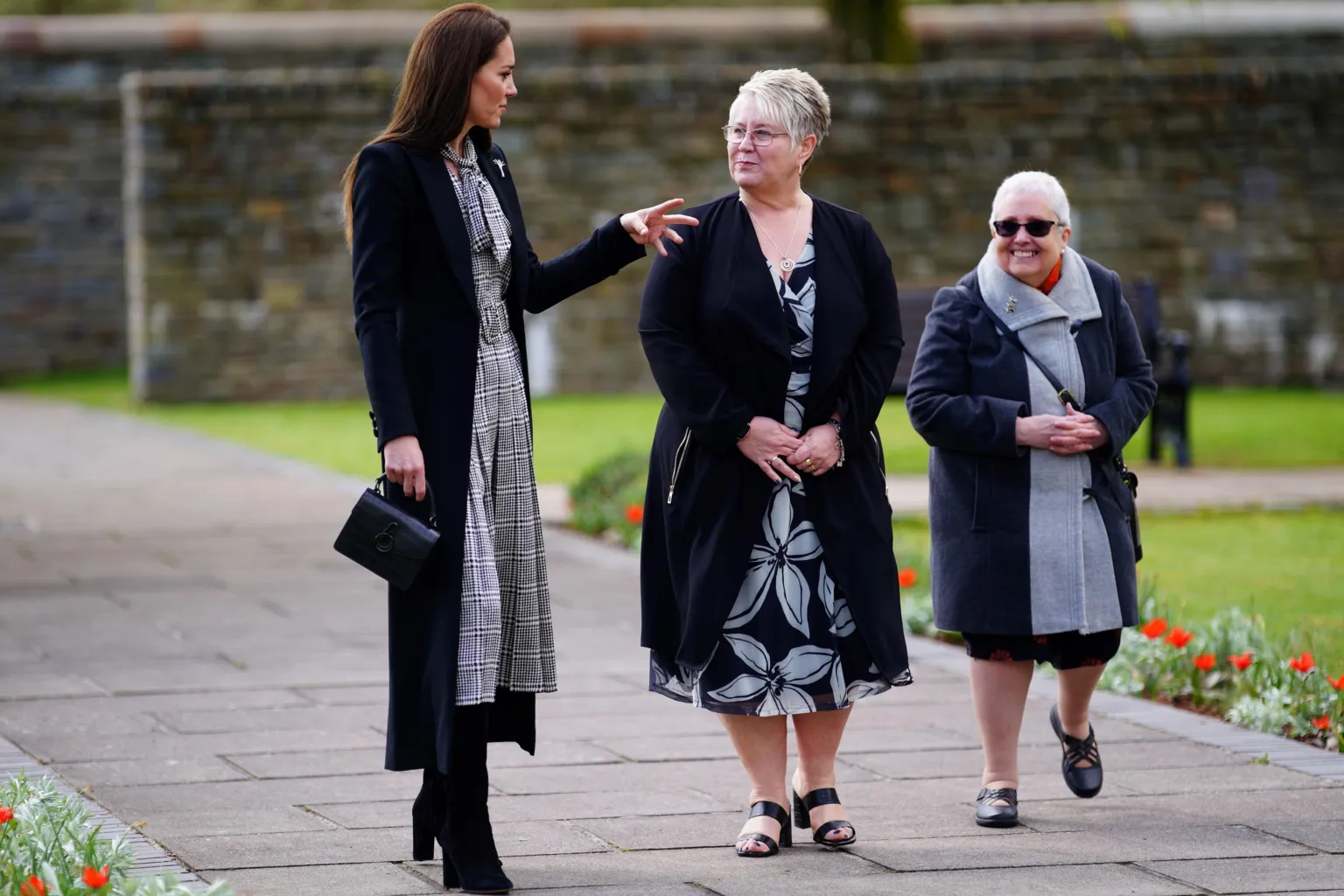  The Princess of Wales speaking to two women in the memorial garden at Aberfan. A neatly-mown lawn and beds with red flowers can be seen in soft focus behind and to the right of the group. They are walking along a paving slab path, with a large stone wall in the background.