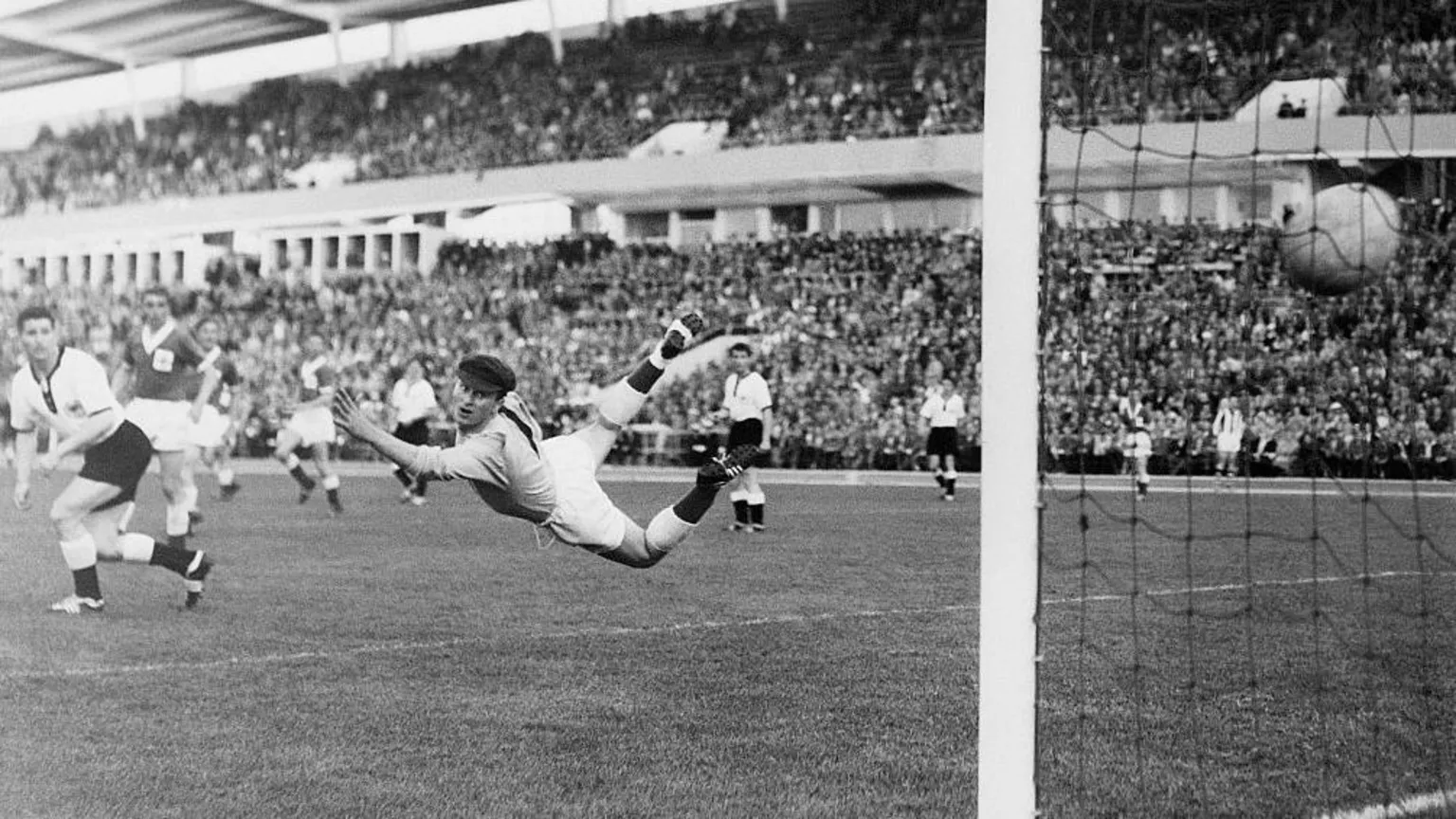  A black and white photo from the 1958 World Cup showing Northern Ireland's goalkeeper Harry Gregg wearing a cap but no gloves diving to try to stop a shot from a German player who is not in shot. He is diving in mid air. The ball has flown past him and can be seen hitting the back of the net at the far right of the image. The game is being played in a stadium packed full of fans.