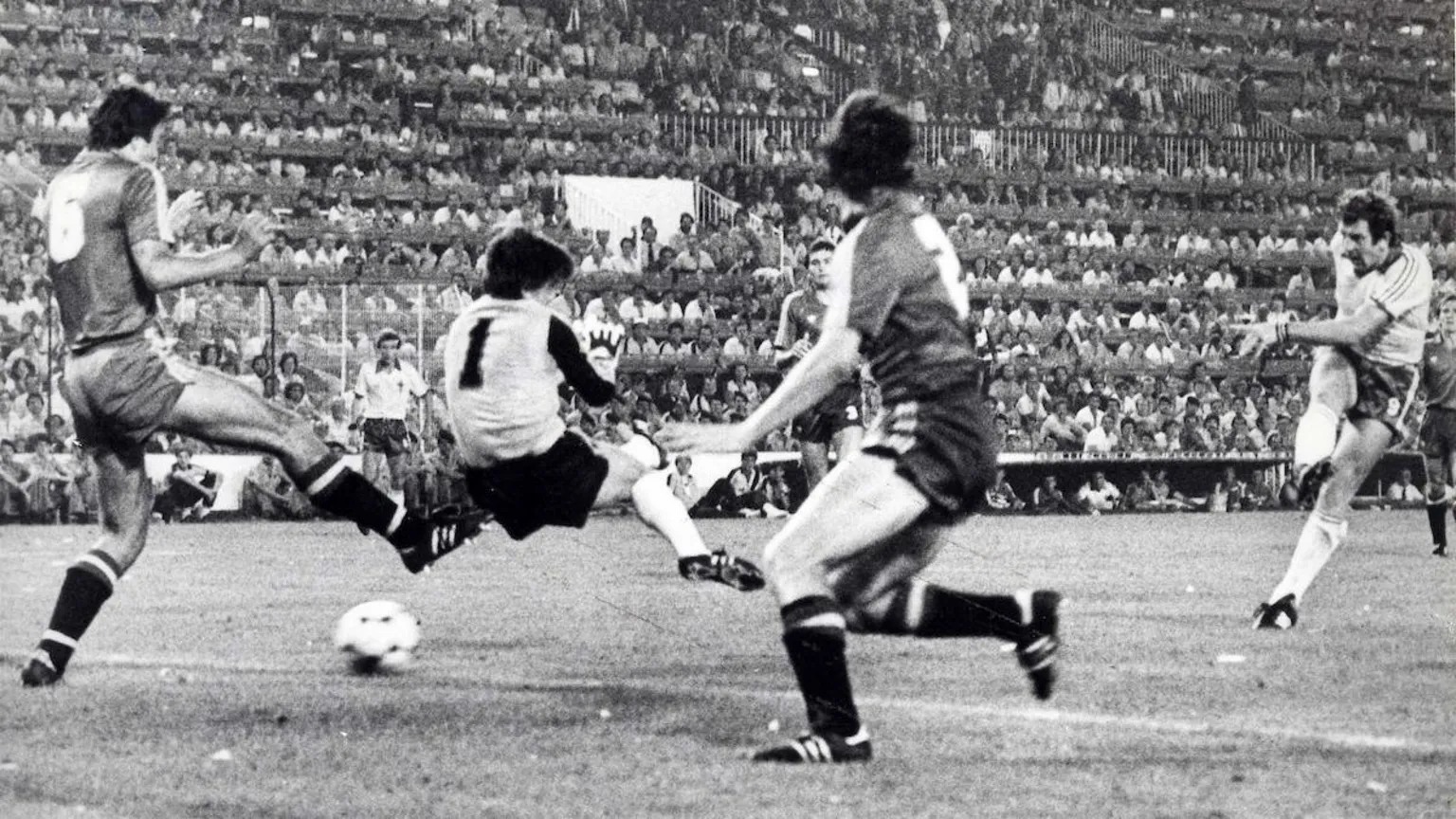  A black and white image of Gerry Armstrong scoring against Spain in 1982. Armstrong is on the far right of the image having taken a shot, he is wearing a white football shirt, dark shorts and white socks. The ball is seen going under the goalkeeper who has black hair and is wearing a light jersey with 