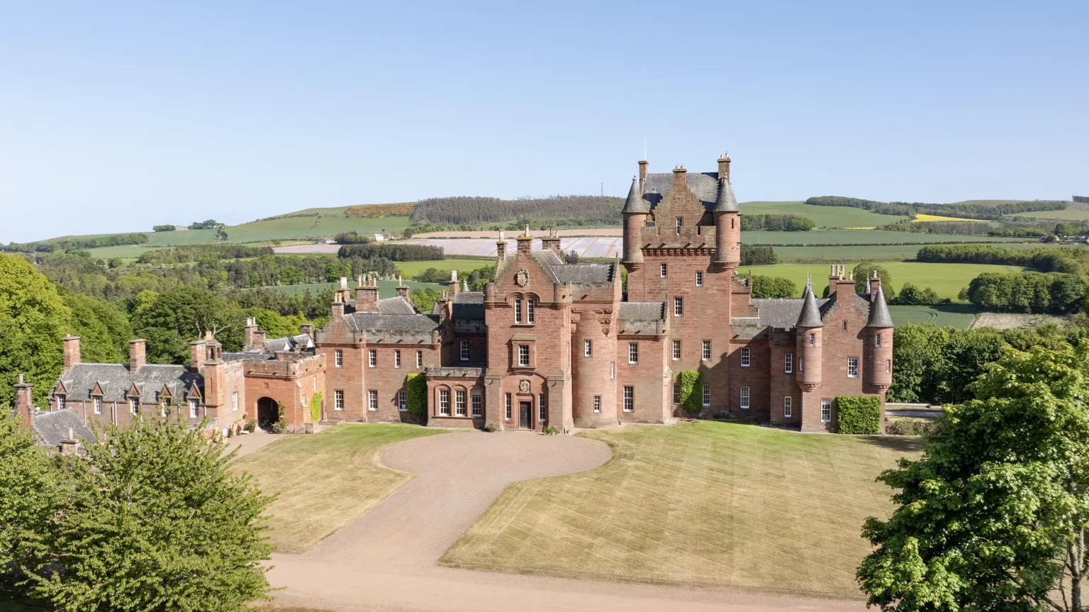 GSC A redstone castle with prominent turrets with fields and trees around it. There is a lawn and a driveway and turning area in front of the main entrance 
